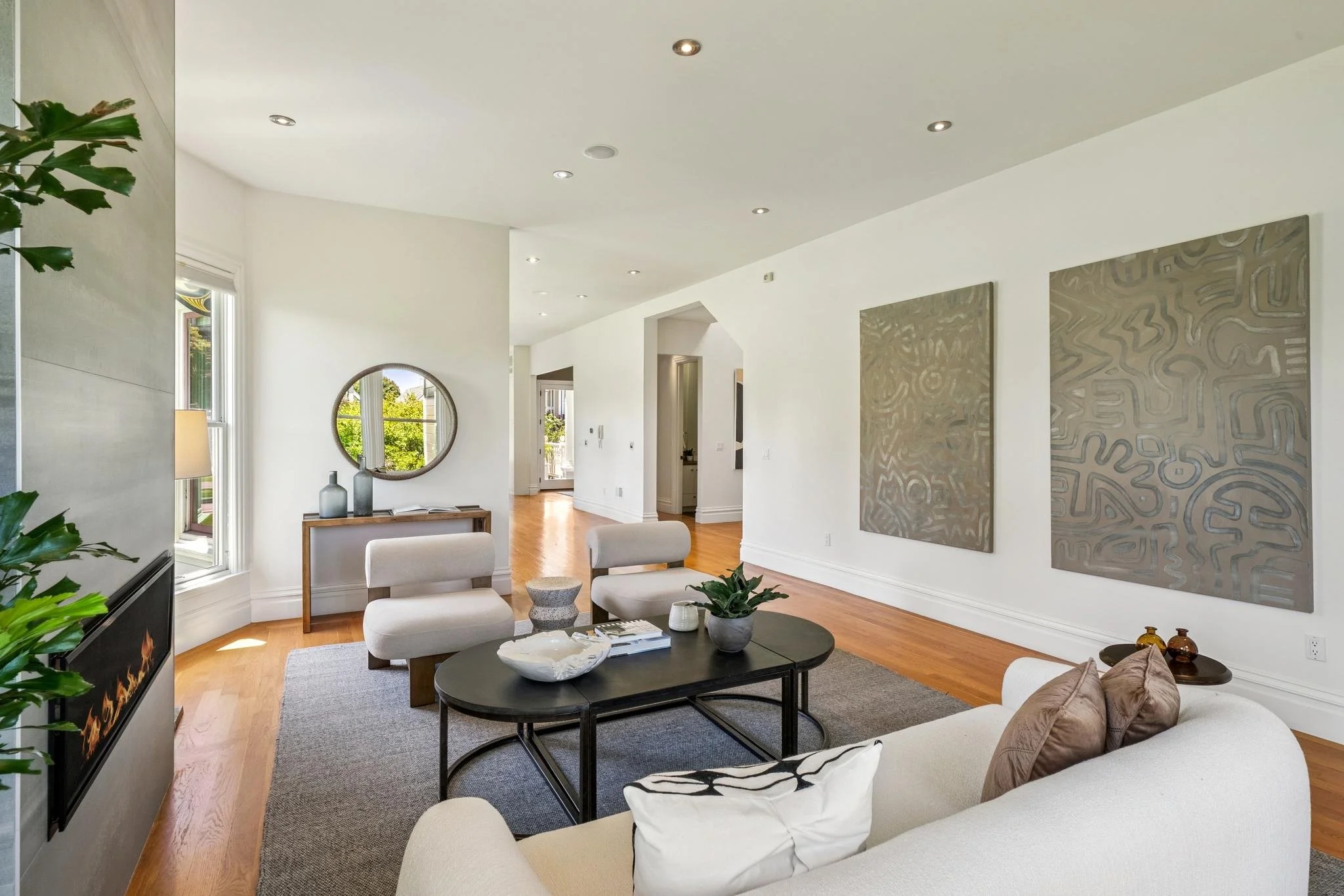 Modern living room with cream sofas, black coffee table, wall art, round mirror, and wooden flooring, bright with natural light from large windows.