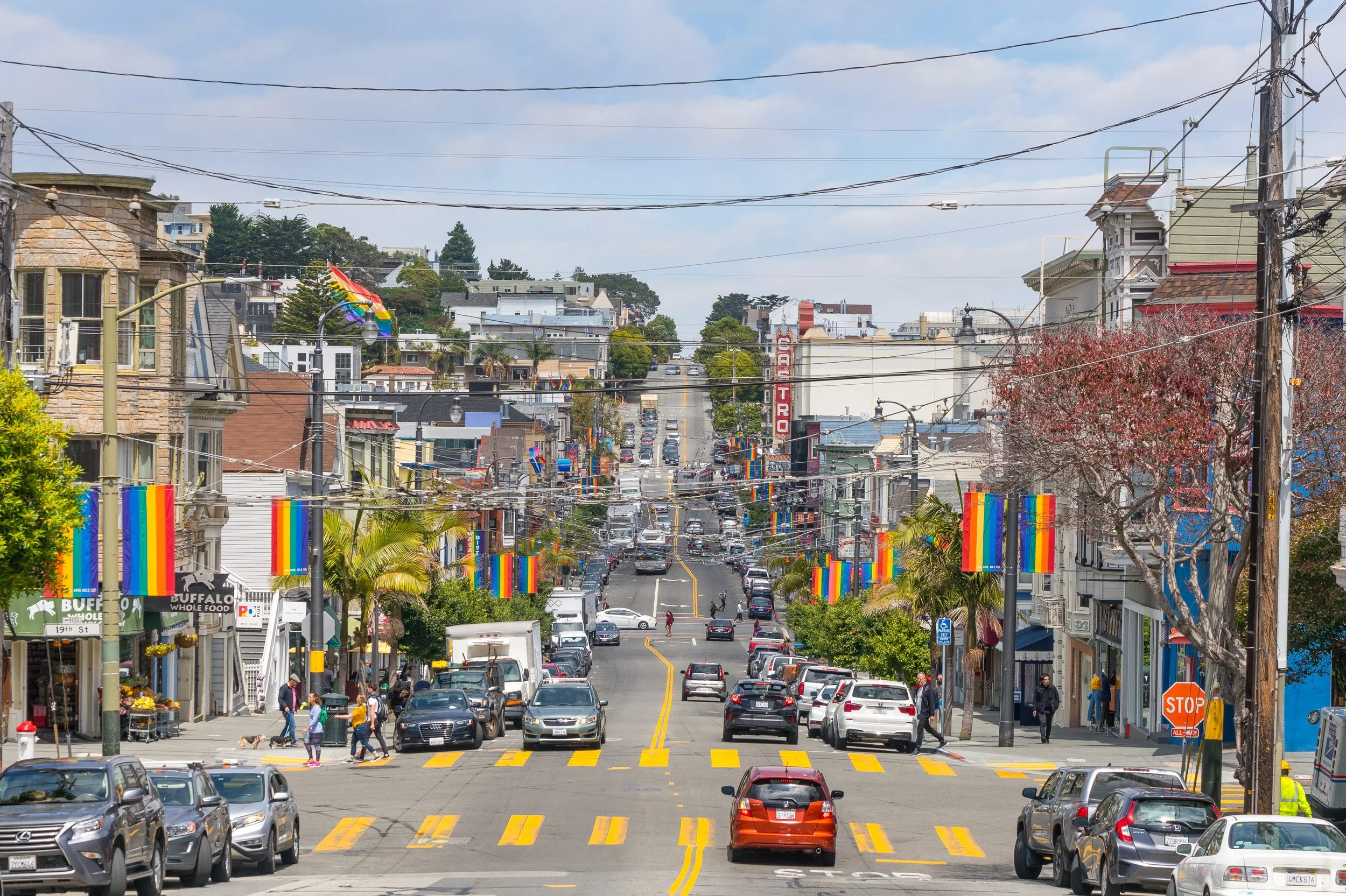 A busy street scene in an urban area with shops, colorful pride flags hanging along the street, cars parked and driving, with people walking on the sidewalks, and a partly cloudy sky overhead.