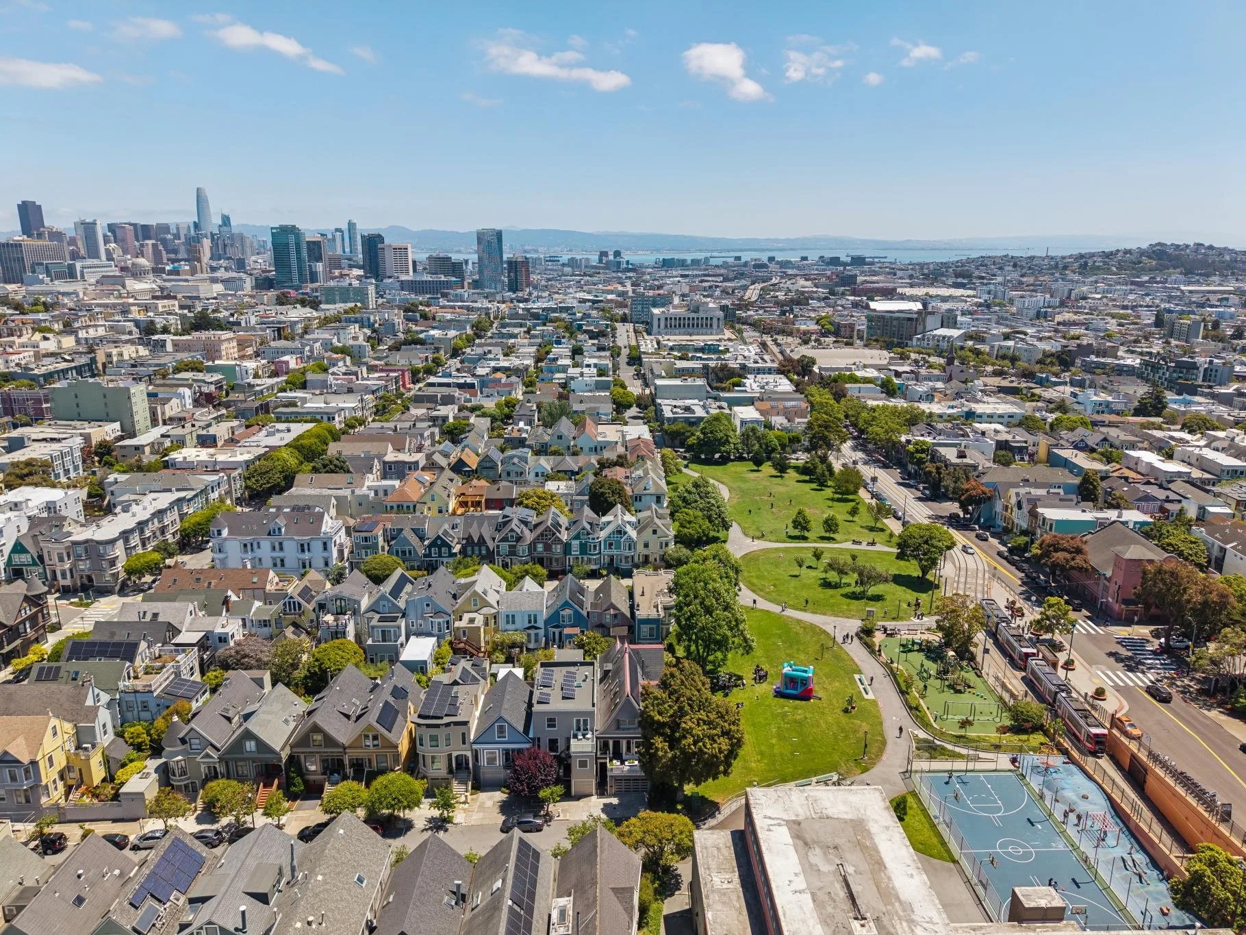 Aerial view of a city with a mix of residential houses, a park with green grass and trees, and a downtown skyline with tall buildings in the distance, under a blue sky with some scattered clouds.