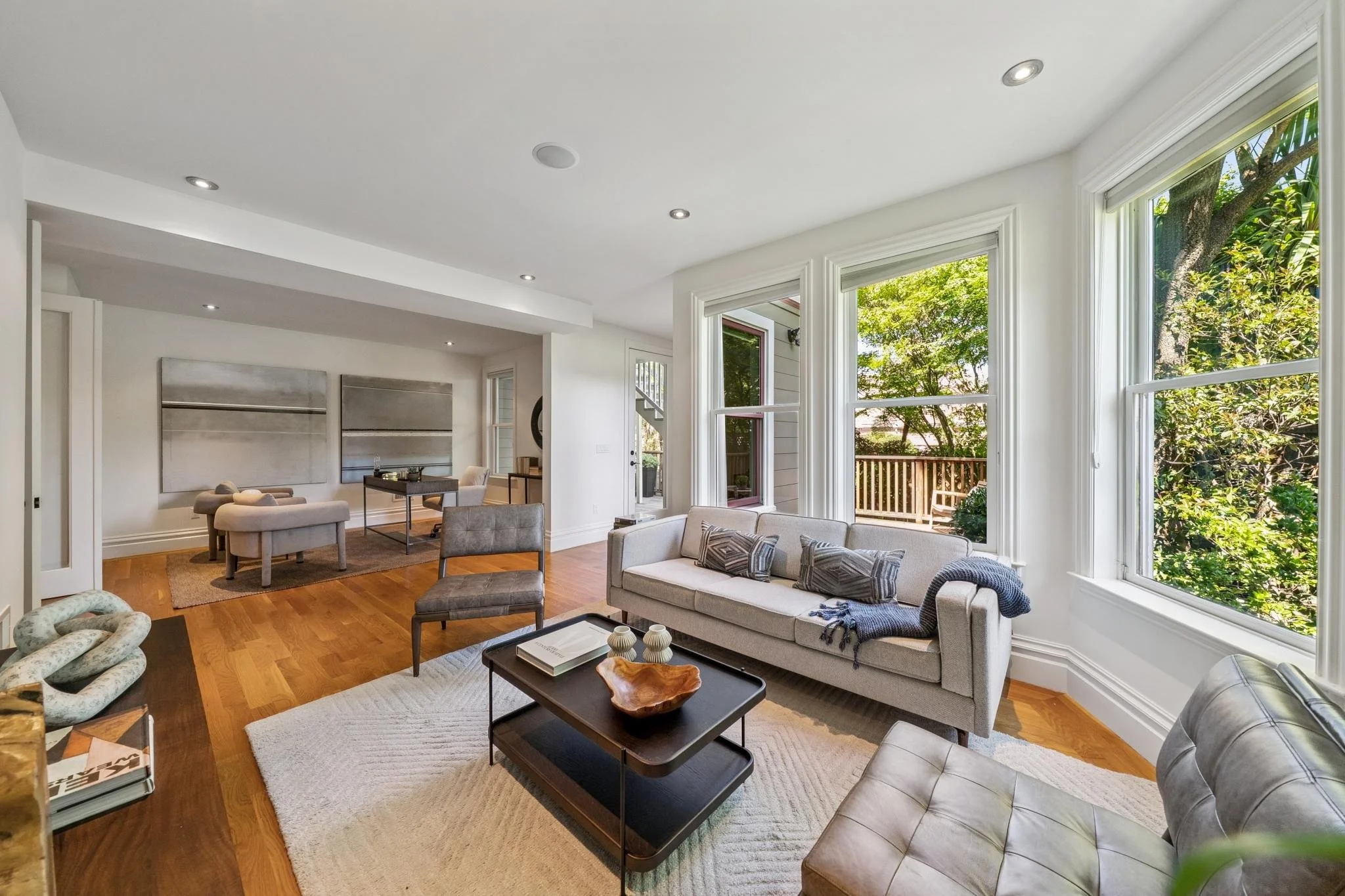 Living room with large windows, white walls, hardwood floors, and modern furniture including sofas, armchairs, and a coffee table with decorative items, overlooking a backyard with trees.