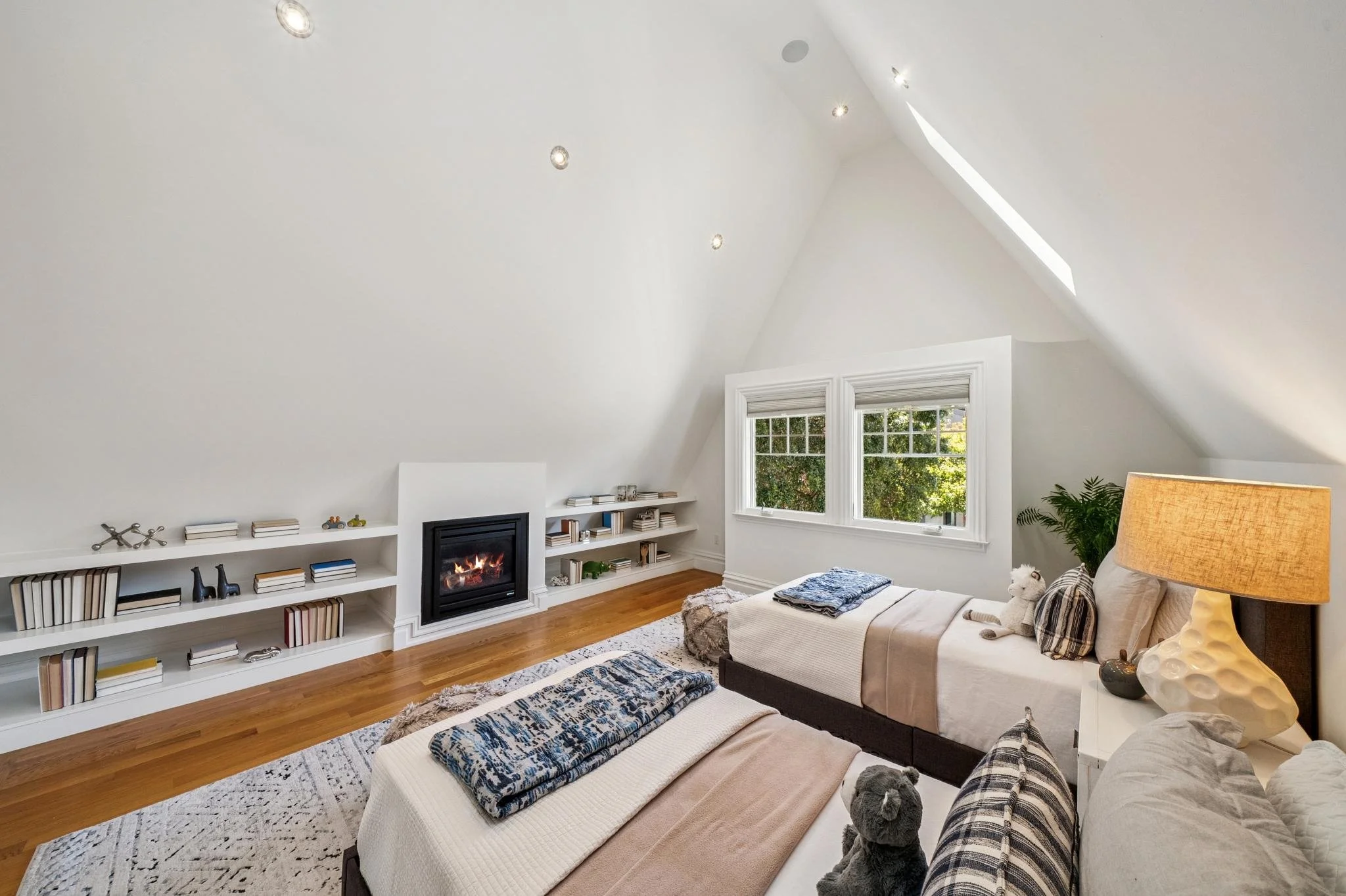 Living room with white painted walls, large windows, a beige sofa with pillows, a table lamp, a white bookshelf filled with books, a fireplace, and decorative items, with hardwood flooring and a patterned rug.