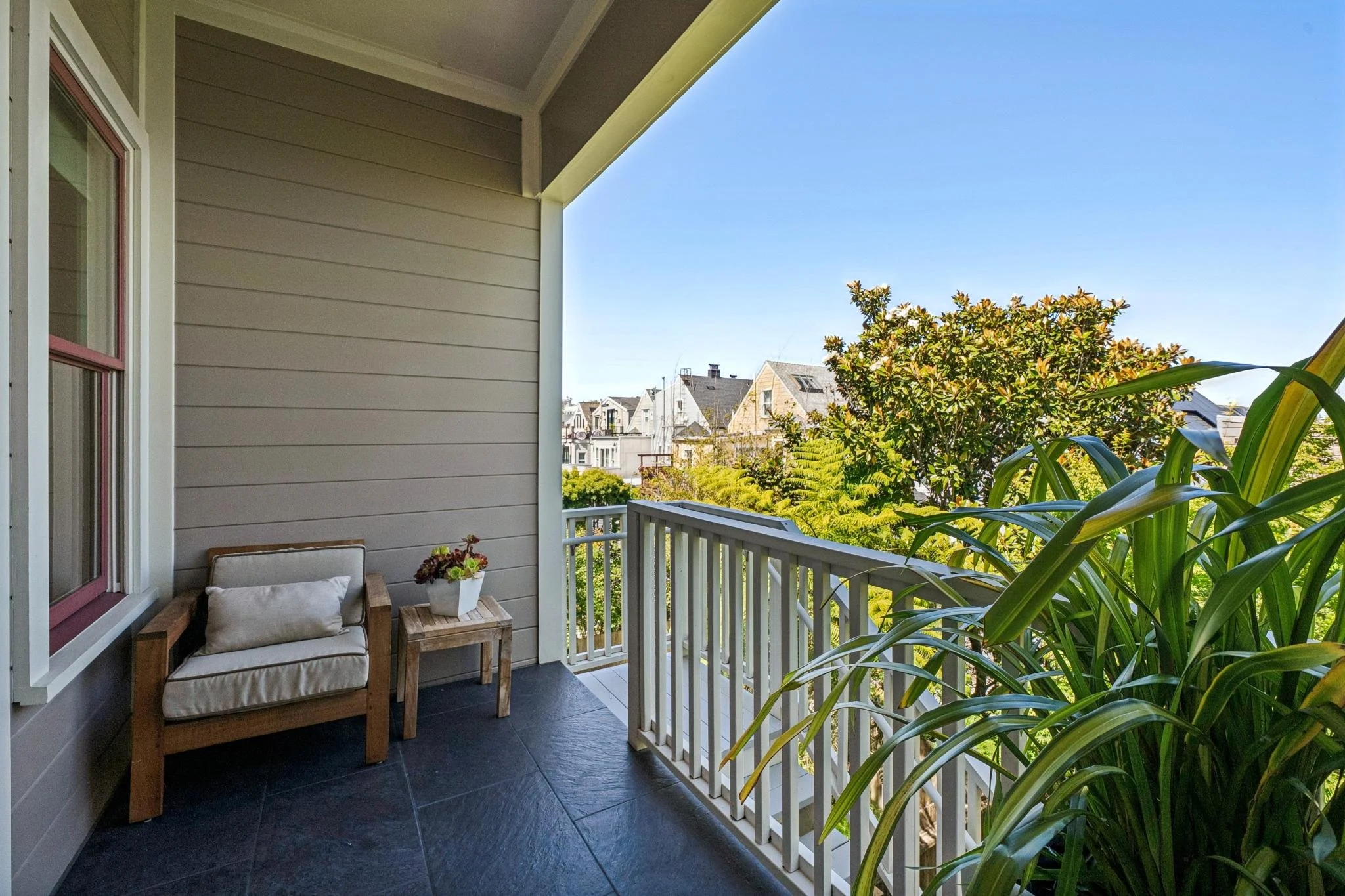 A cozy balcony with a small armchair, a side table with a flowerpot, and lush green plants, overlooking neighboring houses and clear blue sky.