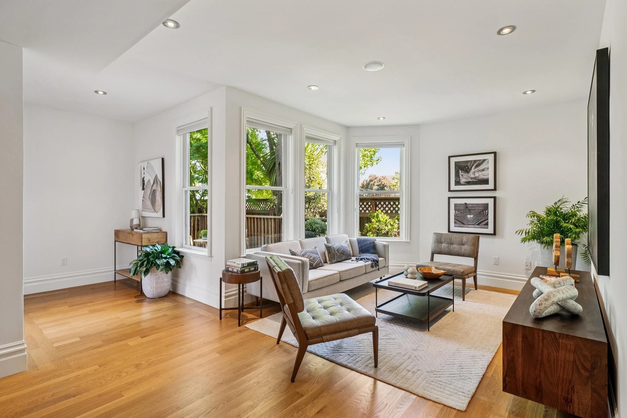 Bright living room with large windows, white walls, hardwood floor, beige sofas, armchair, coffee table, black and white framed artwork, green potted plants, and decorative items.