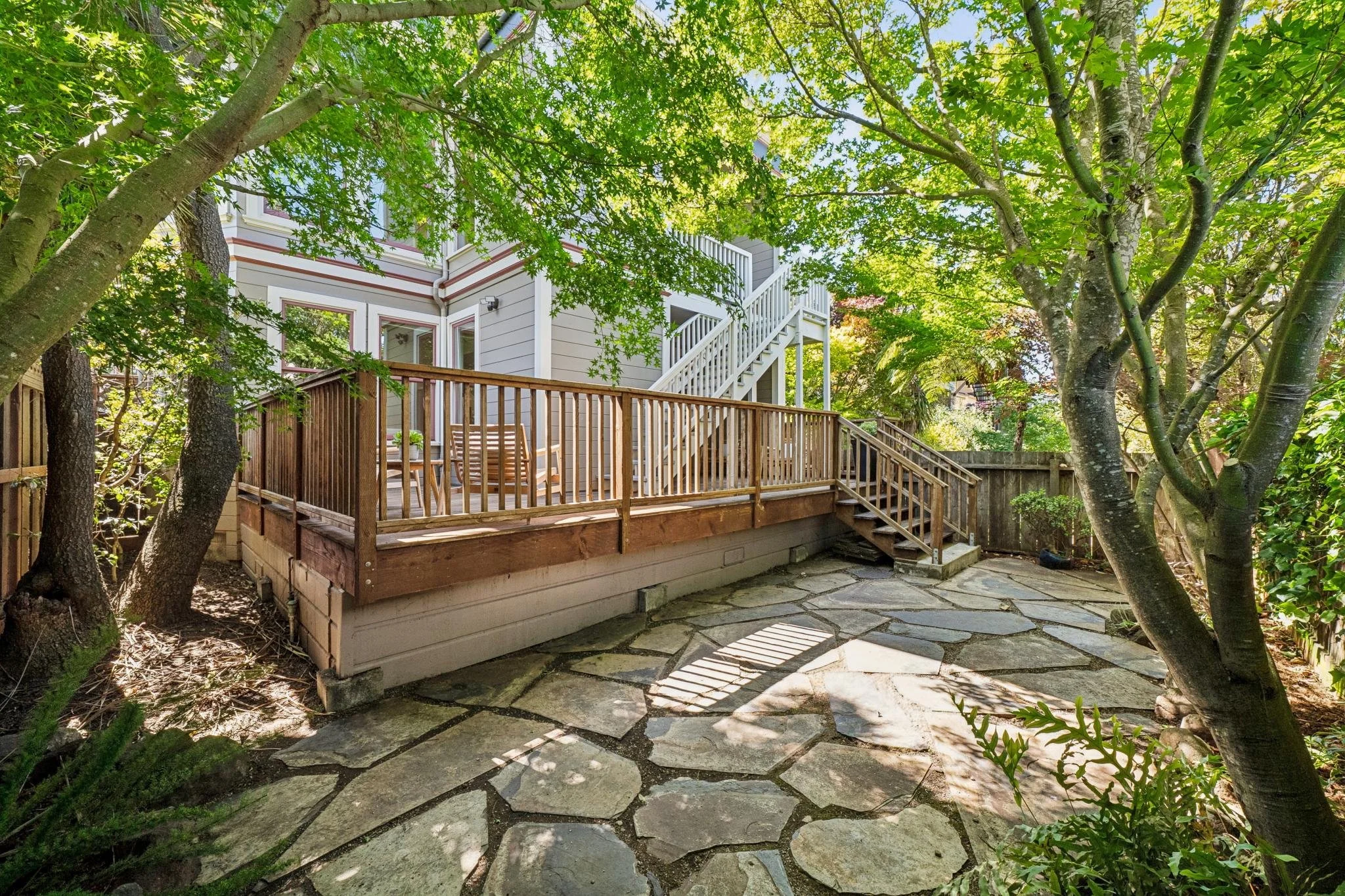 A backyard scene with trees, a wooden deck with outdoor chairs, and a house with stairs visible through the greenery.