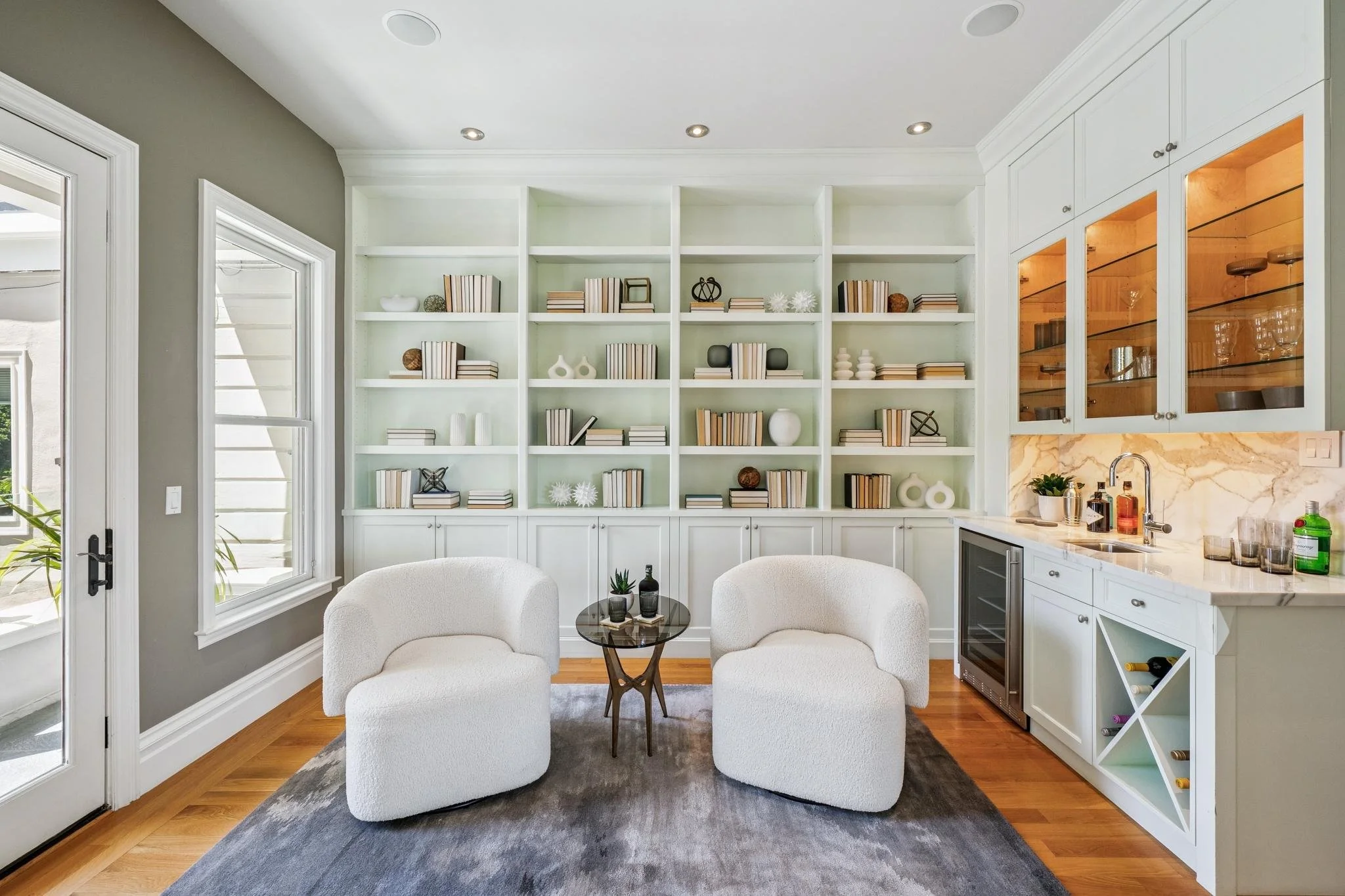 Living room with white built-in bookshelves filled with books and decorative items, two white armchairs, and a small round glass table with potted plants, adjacent to a kitchen with white cabinets, marble countertops, and glass-fronted wooden cabinet