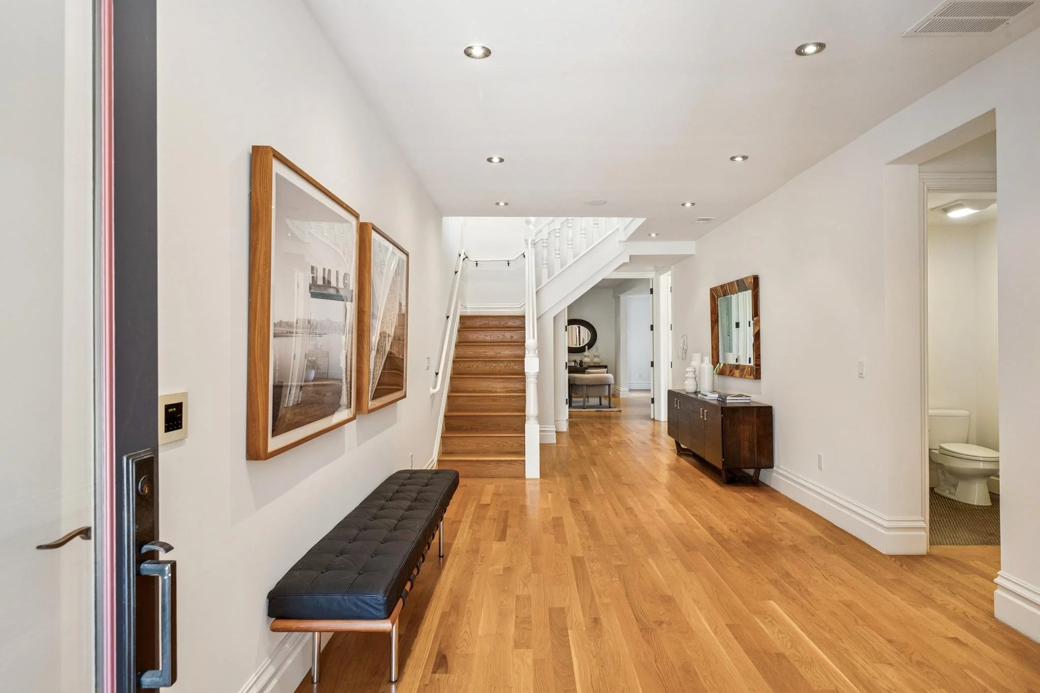 A bright hallway with wooden flooring, white walls, ceiling lights, and a staircase. Decor includes framed pictures, a mirror, a black bench, and a wooden sideboard. A bathroom is visible at the end.