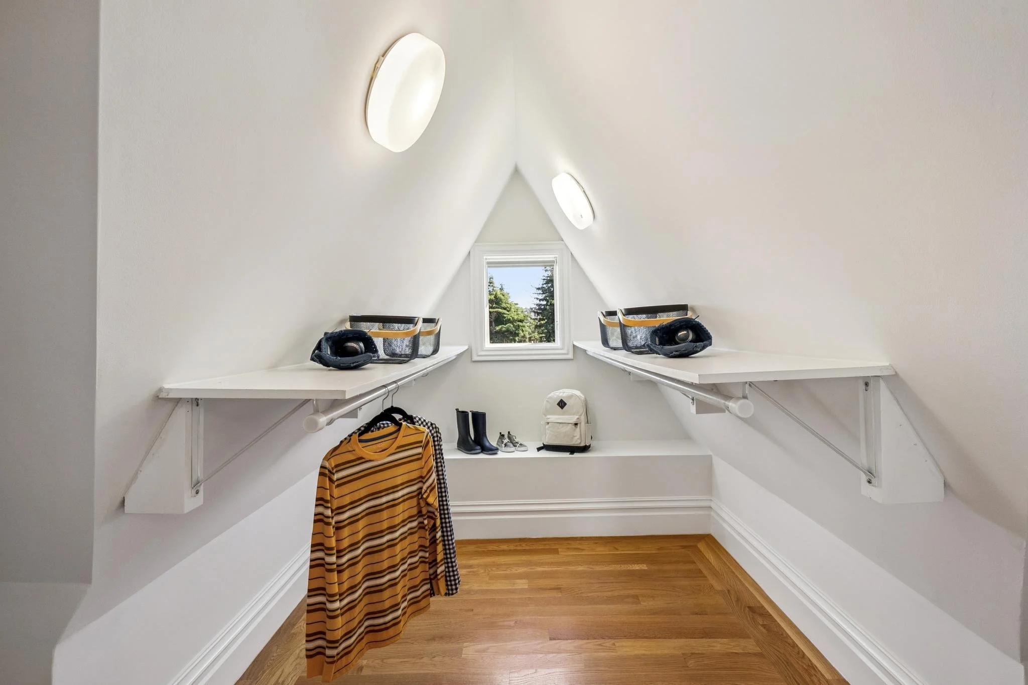 A small white attic closet with sloped ceiling walls, a window with trees outside, and organized shelves with storage baskets and shoes.