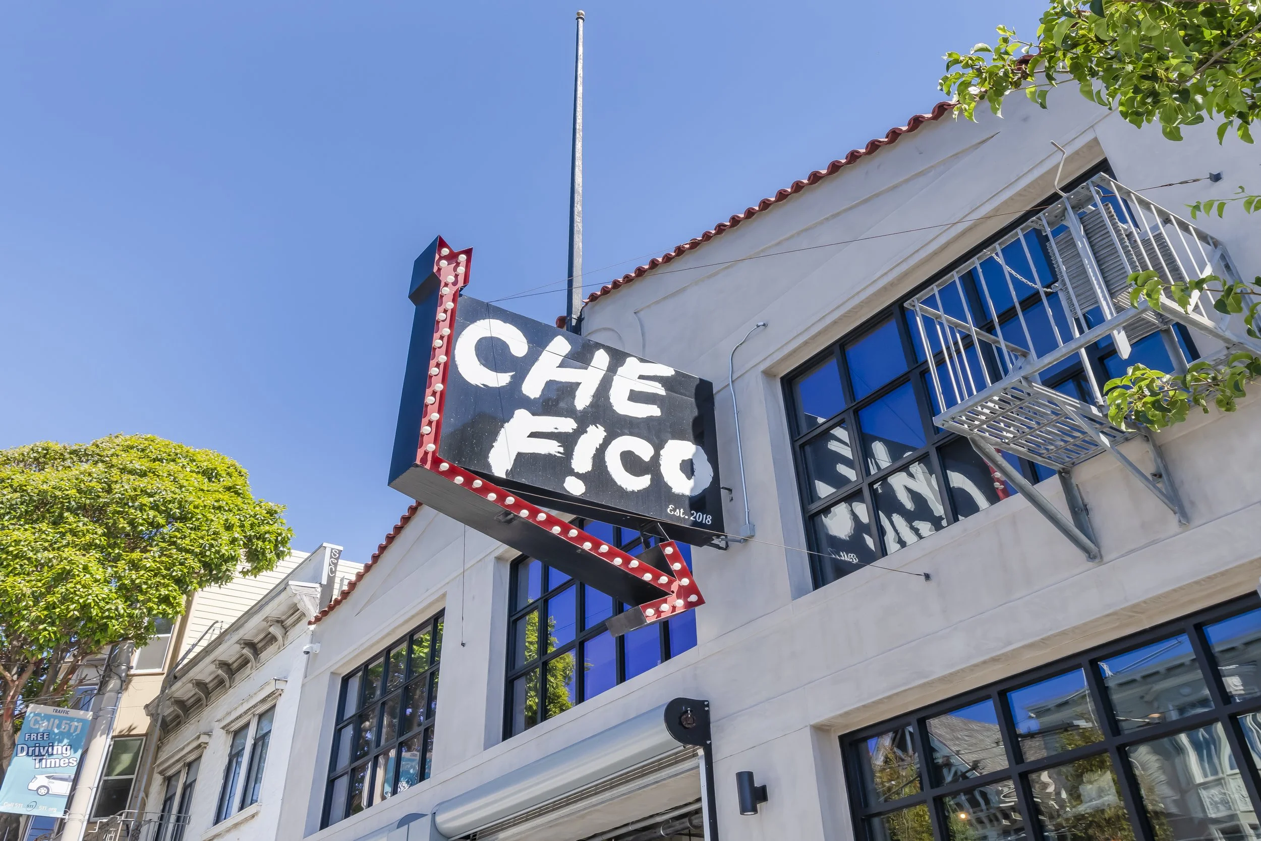 A building with a sign that says 'The Fritz' with a red arrow and light bulbs around its border. The building has a white facade with large black-framed windows, and there is a fire escape on the side. The sky is clear and blue, and there are green t