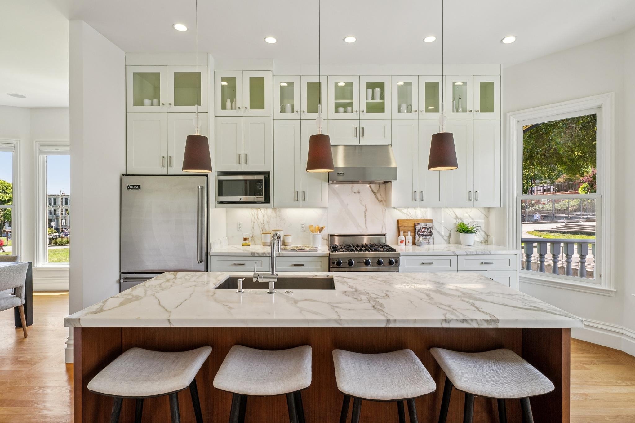 Modern kitchen with white cabinetry, marble countertops, and a large island with four beige chairs. There are pendant lights hanging over the island, and a window showing trees outside.