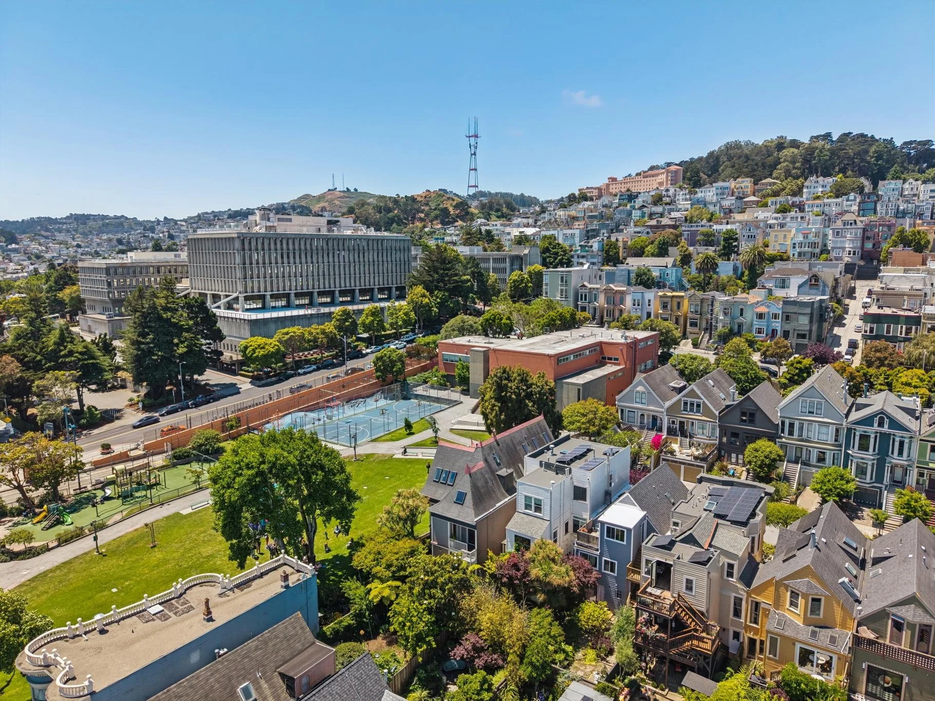 A cityscape with a variety of residential buildings, a sports court, playground, and greenery under a clear blue sky, with hills and telecommunications towers in the background.