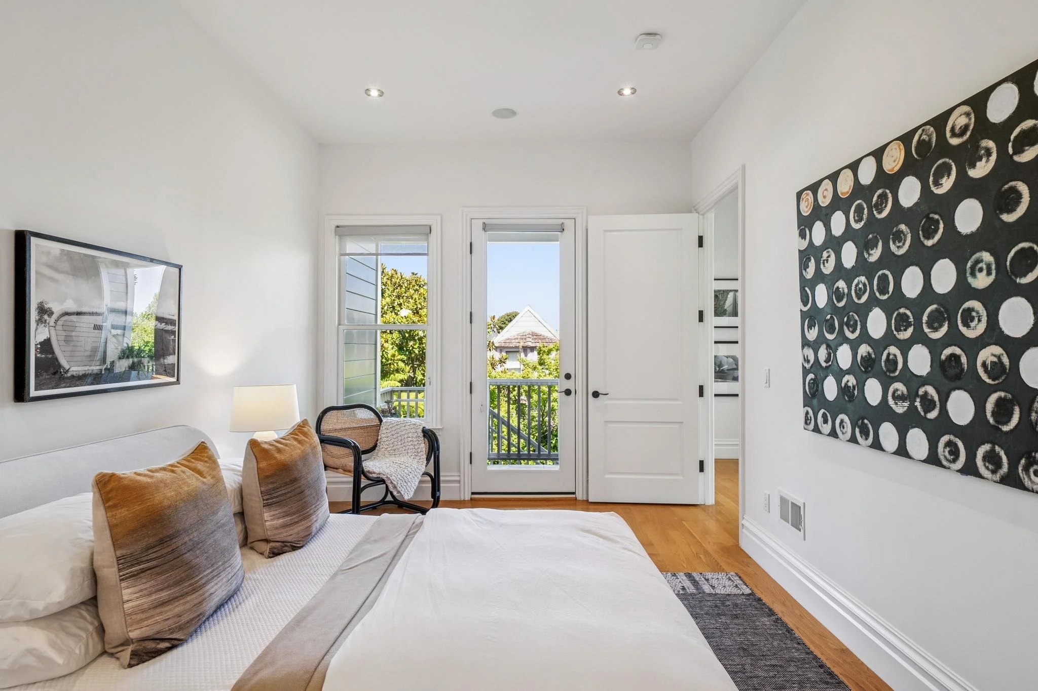 A cozy living room with a white sofa, tan pillows, and a black chair with a throw blanket. Natural light streams through a glass door and window, leading to a balcony with trees and a neighboring house visible outside. The walls feature black and whi