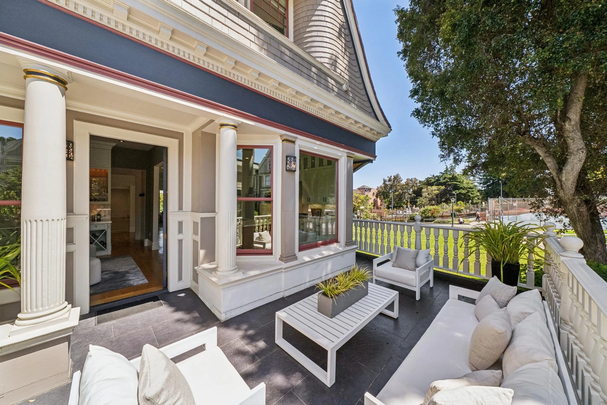 A residential porch with white columns, wicker furniture with beige and white cushions, potted plants, and a black tile floor, overlooking a green lawn and trees.