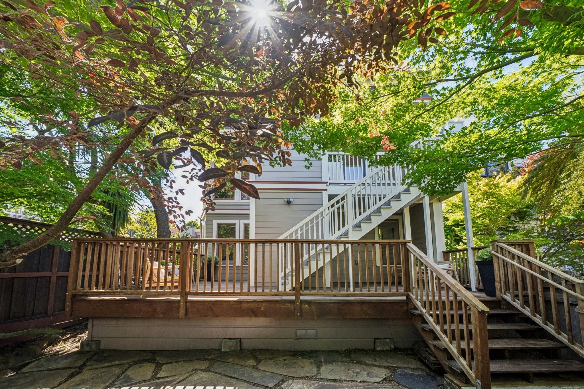 Backyard with a wooden deck, surrounded by trees and foliage, house with a staircase leading to the upper level, sunlight filtering through the leaves.