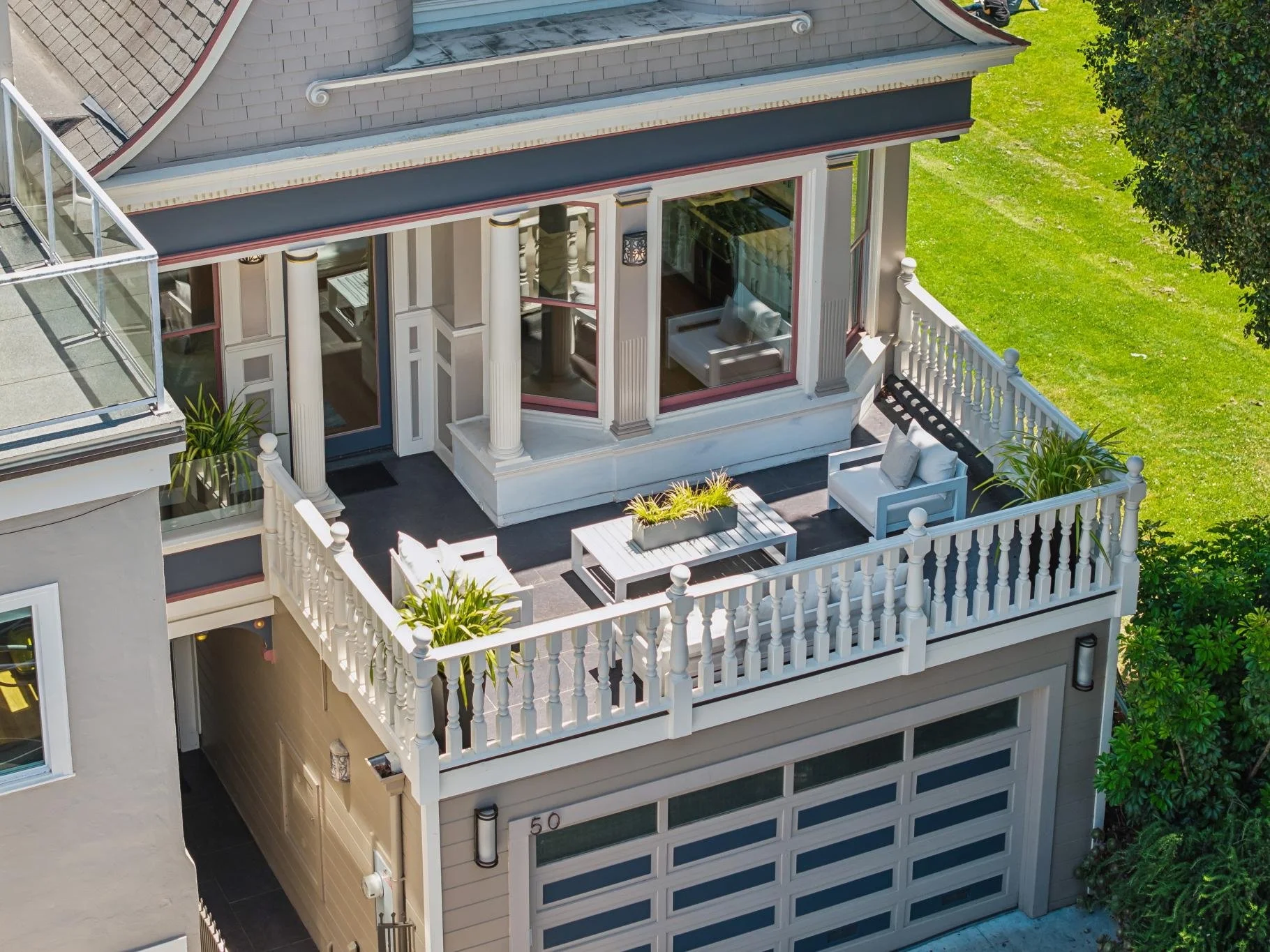 A two-story house with a balcony and a rooftop terrace. The balcony has white railings with decorative posts and outdoor furniture, including a white chair and a coffee table with a plant. The rooftop terrace has a glass railing and a view of the str