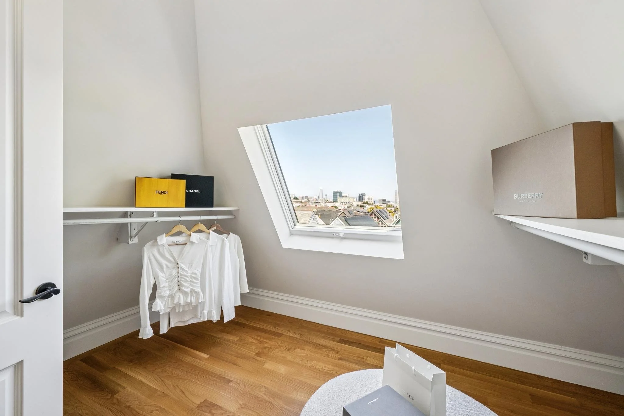 A room with a sloped ceiling and a skylight window showing a cityscape. There are two shelves with shopping bags, one labeled Fendi and the other Chanel, a white shirt hanging on a rack, and a white round rug on a wooden floor.