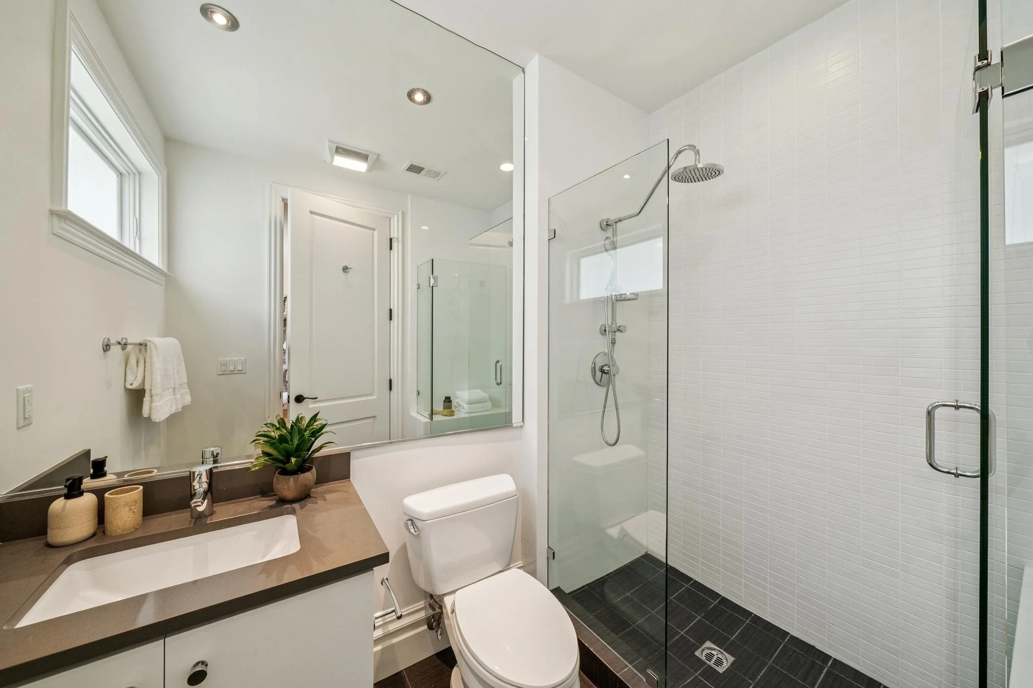 A modern bathroom with a walk-in shower enclosed by glass, white tiled walls, a white toilet, a vanity with a dark countertop, a large mirror, small potted plant, and modern decor in neutral tones. Natural light from a window illuminates the space.