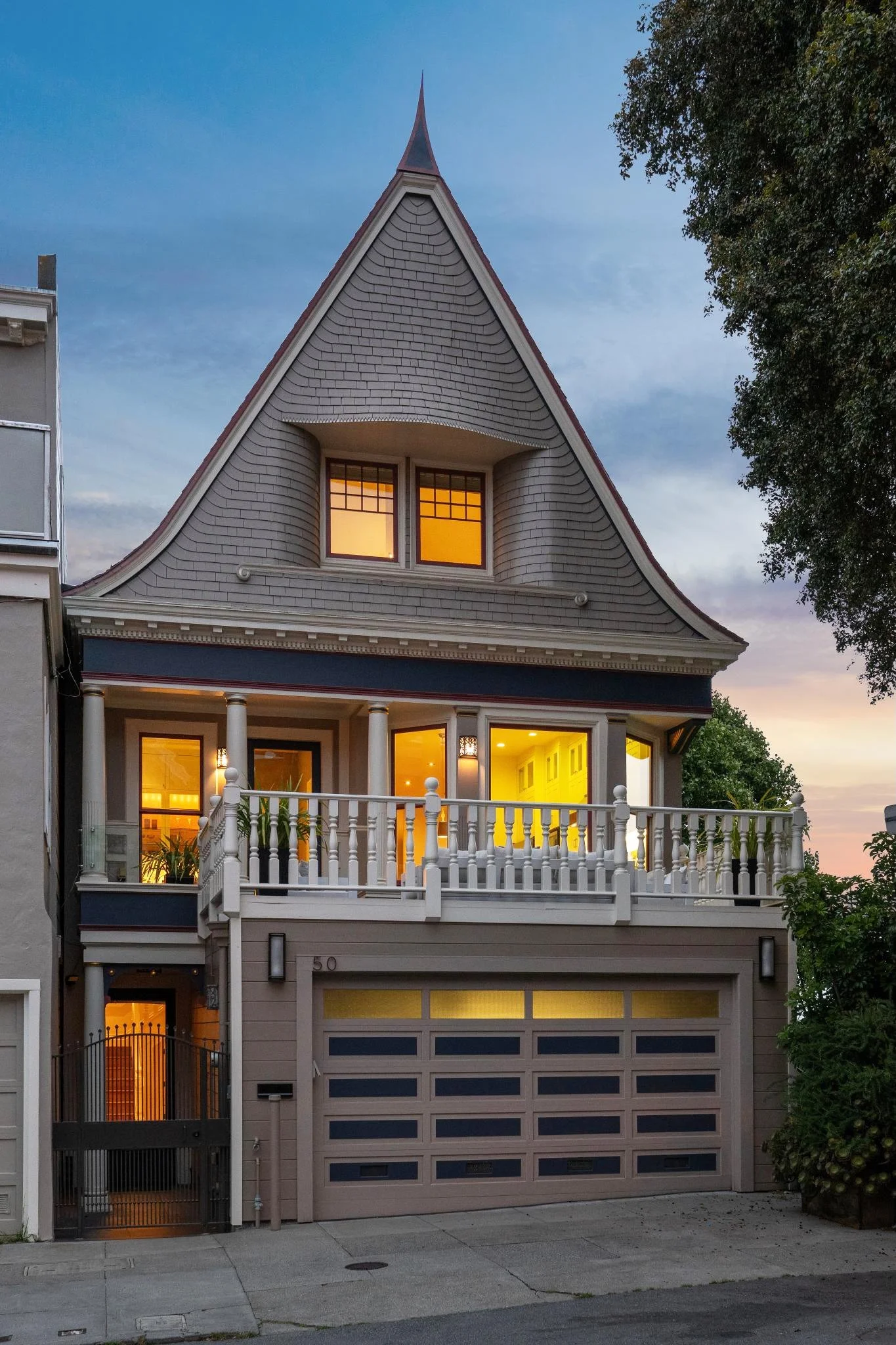 A multi-story house with a steep, pointed roof, gray shingles, and a front porch with white columns and a railing. The house has illuminated interior windows and a gray garage door. There is a tree on the right side and neighboring buildings to the l