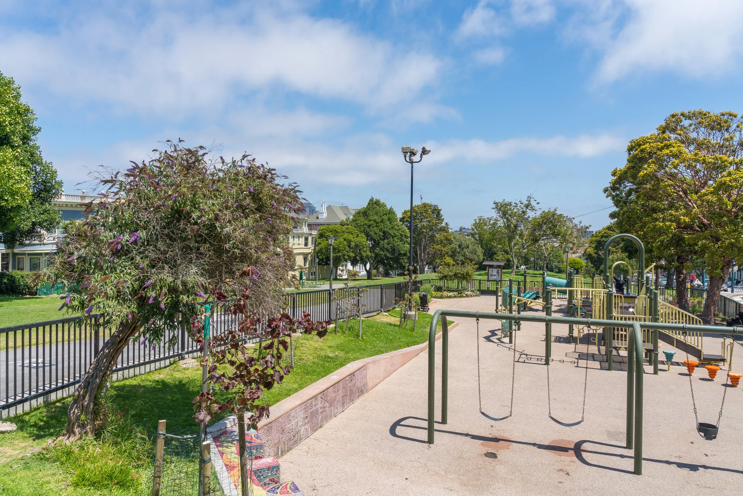 A sunny park with a playground, trees, a grassy area, a fence, and residential buildings in the background.