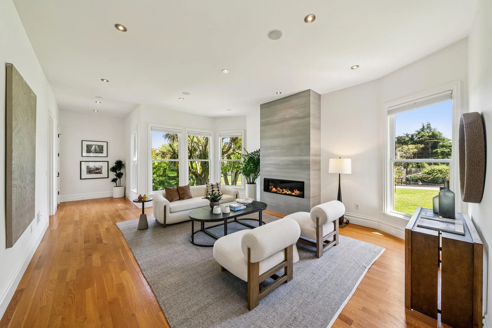 Living room with white walls, hardwood floors, large windows showing trees, a modern fireplace, white sofas, a black coffee table, side tables, and lamps.