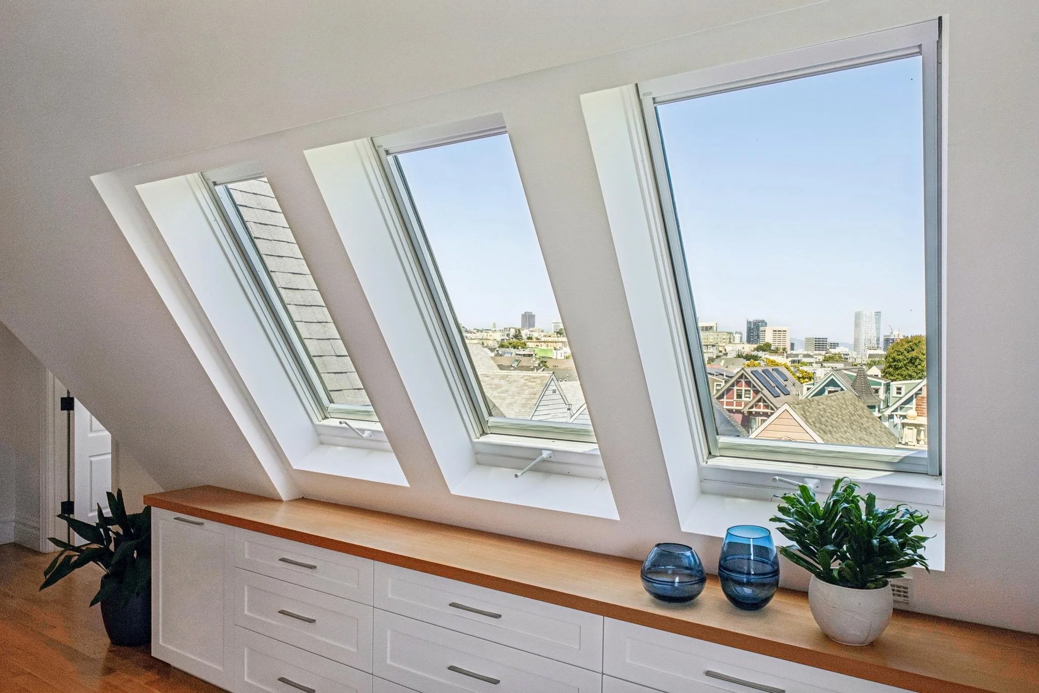 Room with three long skylight windows on a sloped ceiling, view of cityscape and blue sky outside. Interior features a white dresser with wooden top, three blue glass vases, potted plant, and a black speaker.