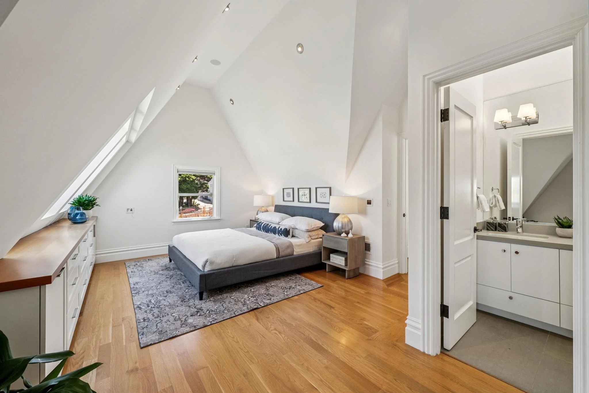 A bedroom with white sloped walls and ceiling, hardwood floors, a gray bed with white bedding and decorative pillows, a wooden dresser with a plant, and a small window showing trees outside. There are bedside lamps and framed artwork above the bed. A