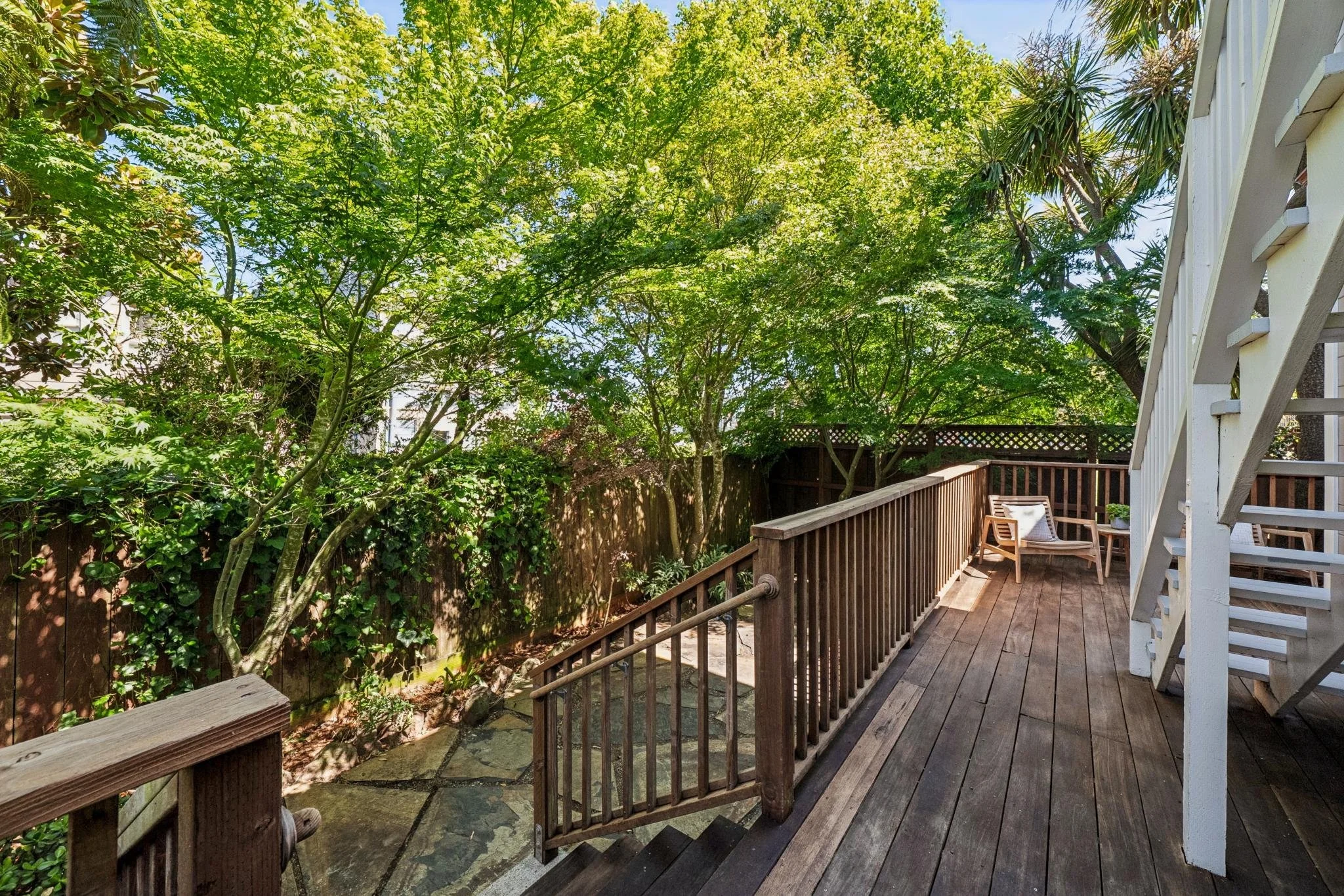 A backyard deck with chairs, surrounded by trees and greenery, with a wooden fence and house exterior visible.