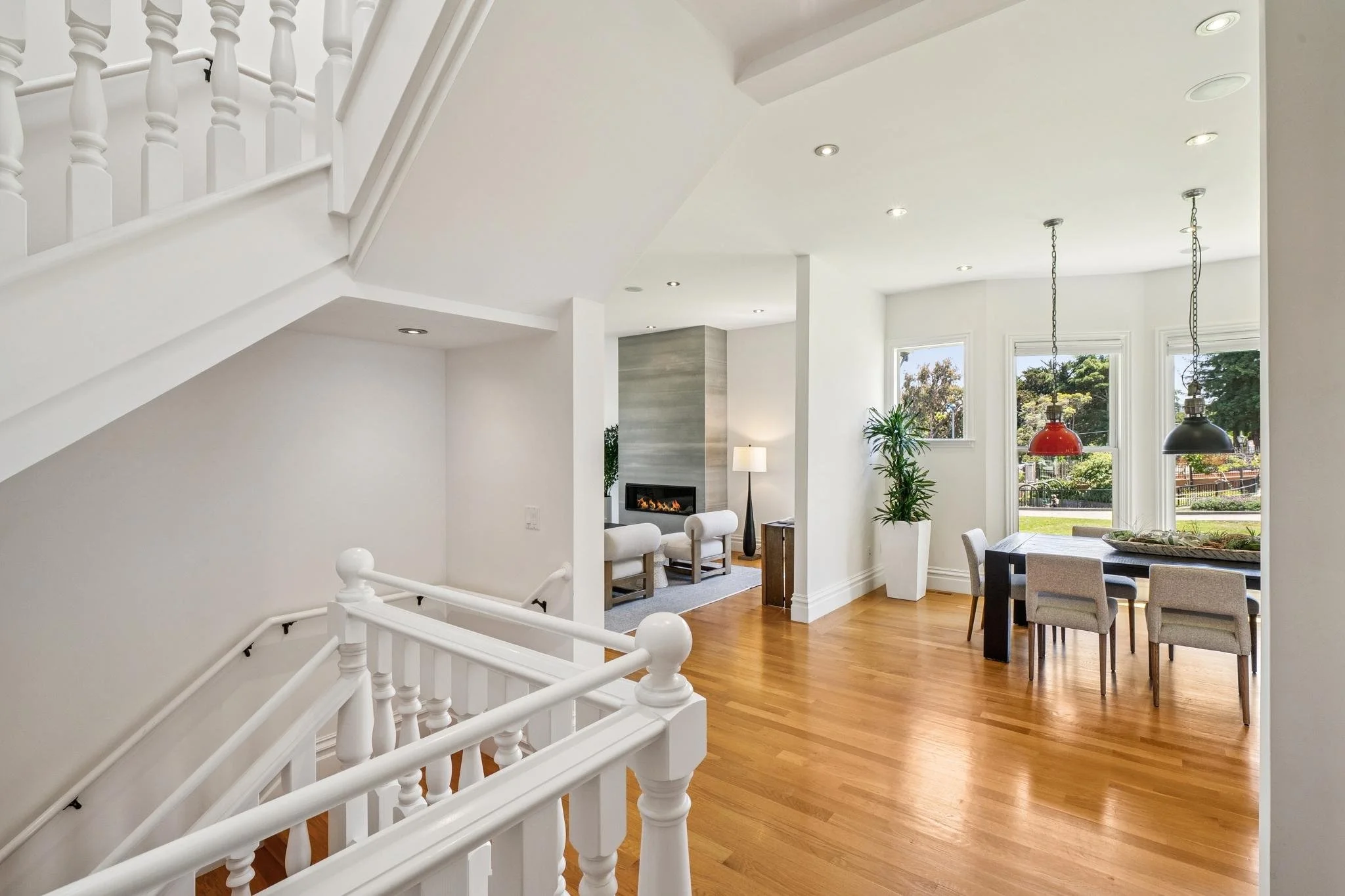 Bright living space with hardwood floors, large windows, and modern decor. Includes a staircase with white railing, some seating, plants, a fireplace, and hanging pendant lights.