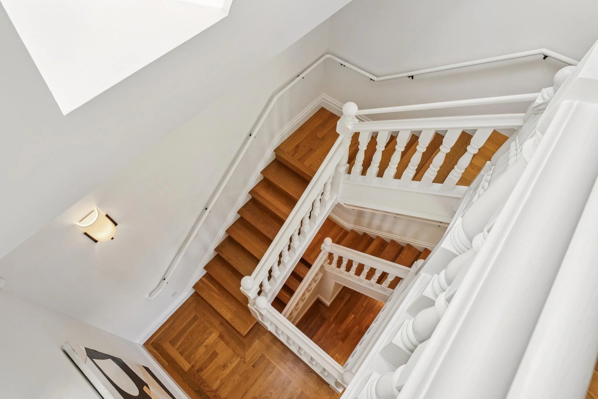 View of a wooden staircase with white balustrades from above, leading down to a lower floor, with a wall-mounted light fixture on the left wall.