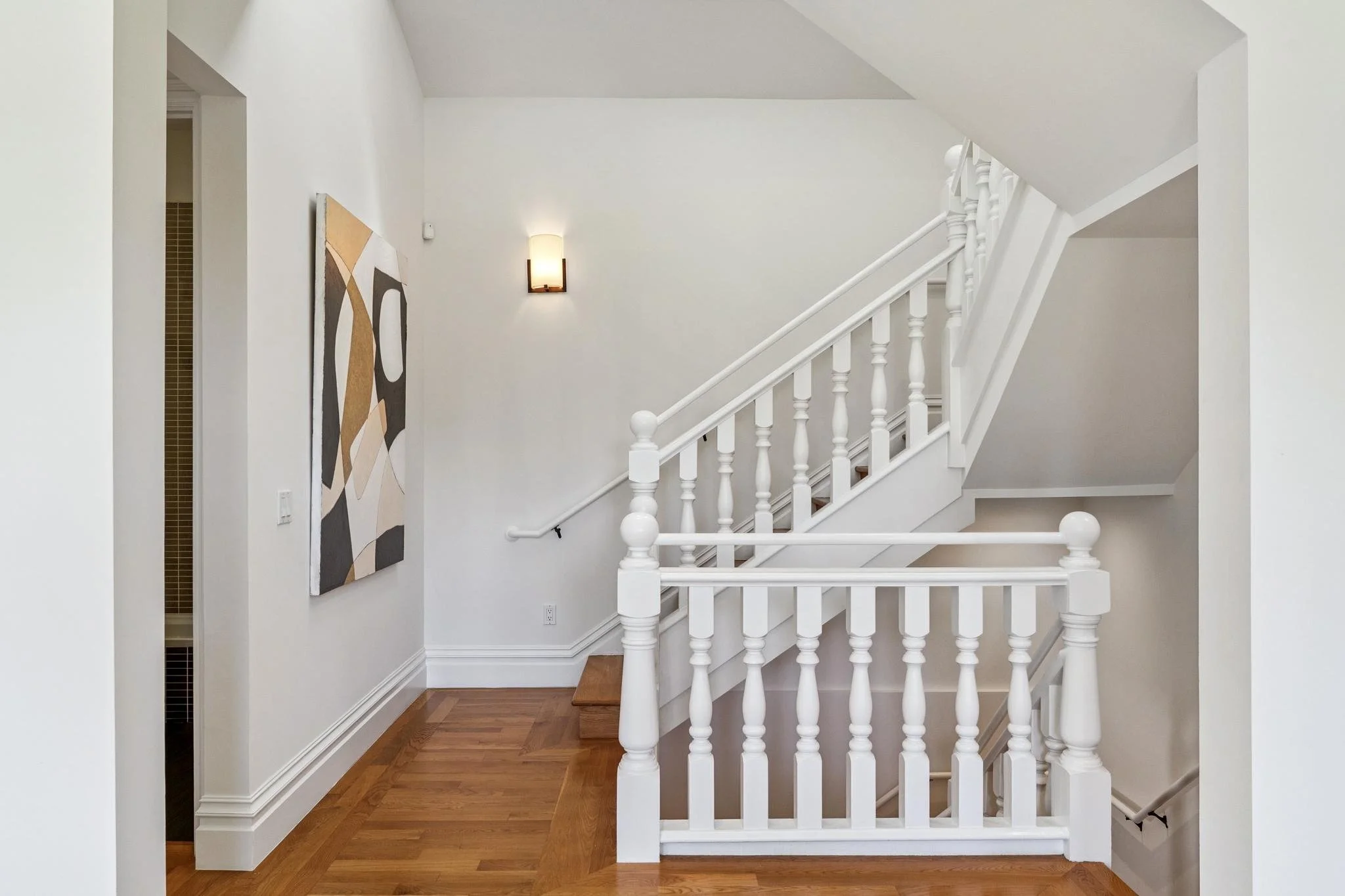 Interior staircase landing with white railing, wooden floors, abstract wall art, and a wall-mounted light fixture.