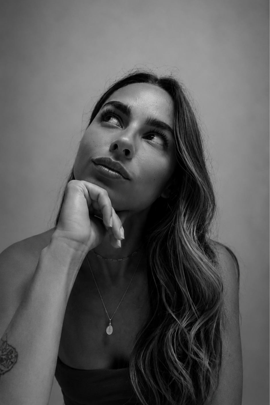 Black and white photo of a woman with long wavy hair, resting her chin on her hand, looking up and to the side with a thoughtful expression. She is wearing a necklace and a sleeveless top.