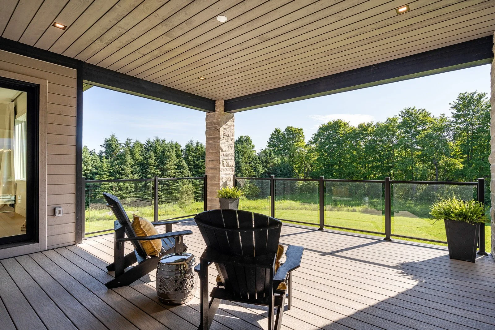 View of a spacious outdoor balcony with wooden flooring, black Adirondack chairs with yellow cushions, potted plants, and a view of green trees and fields under a clear blue sky.