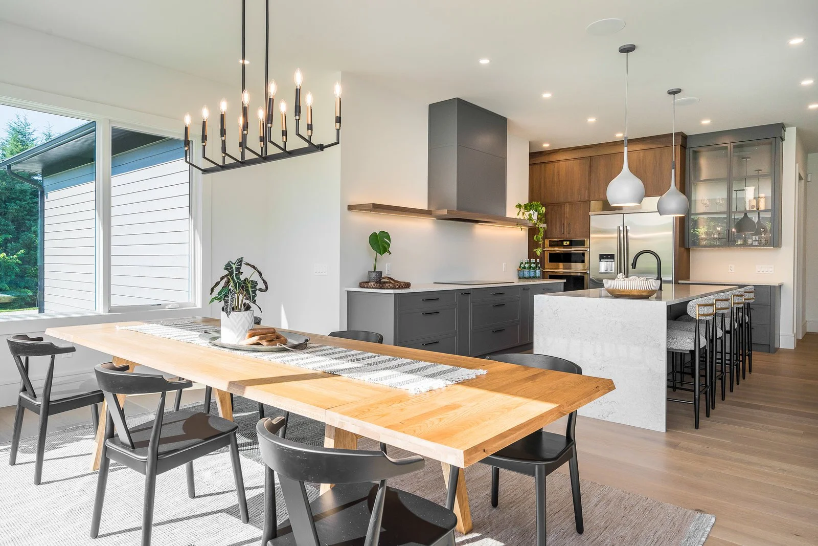 Modern open-concept kitchen and dining area with large window, black chairs, wooden dining table, potted plants, and sleek cabinetry and appliances.