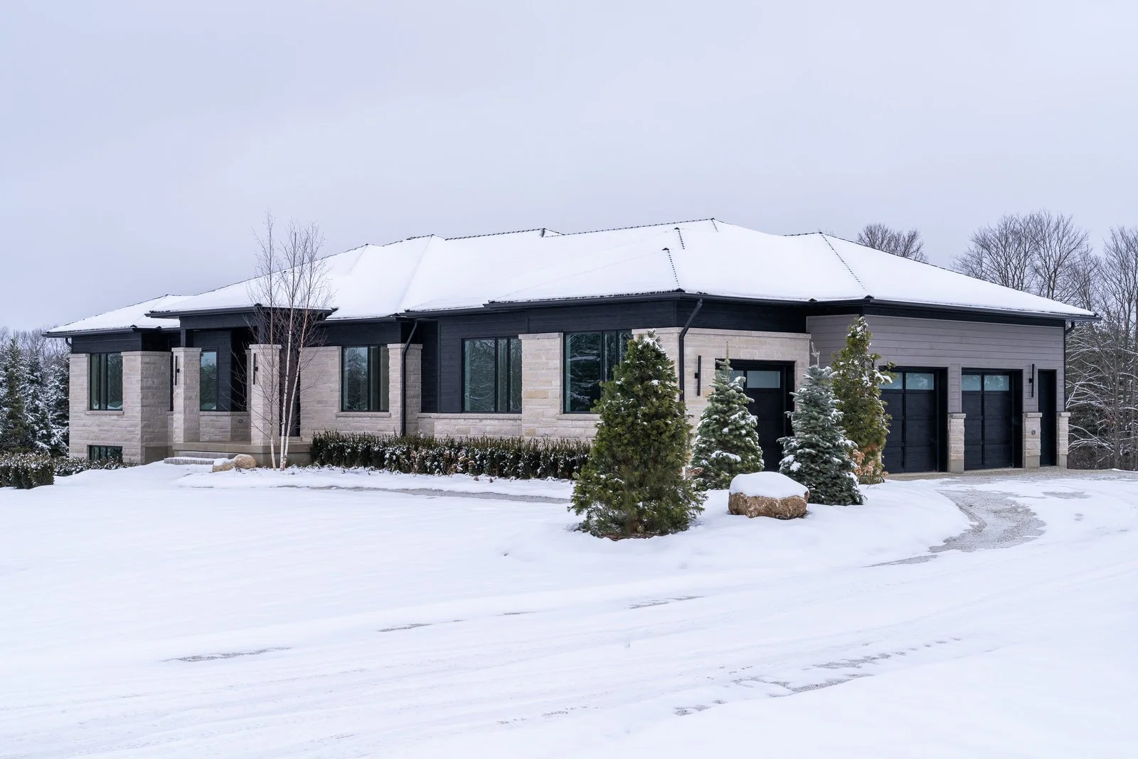 Modern house in winter with snow on the ground and roof, several evergreen trees, a driveway, and a backdrop of leafless trees.