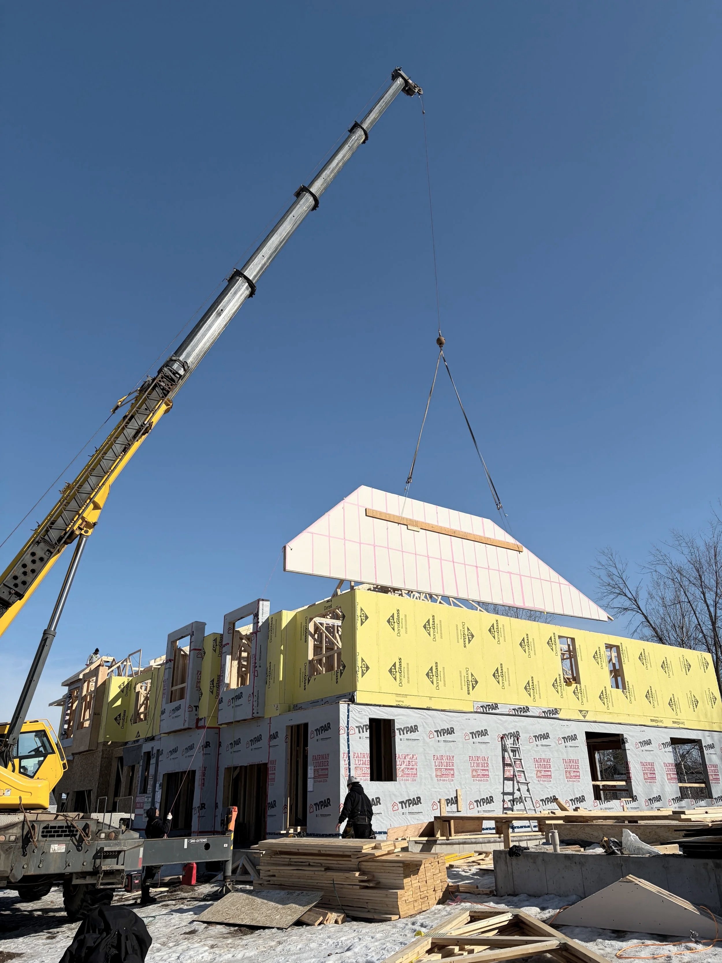Construction site with a multi-story building under construction, workers, wood materials, and a crane lifting a large panel into place under a clear blue sky.