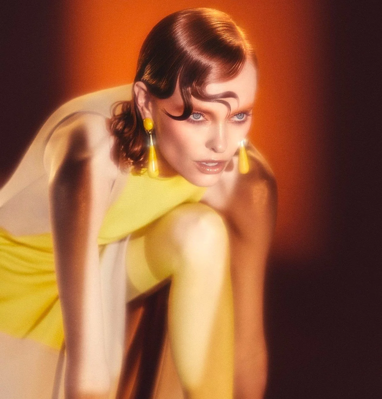 A woman with styled hair wearing yellow earrings and a yellow dress in a crouching pose against a warm orange background.