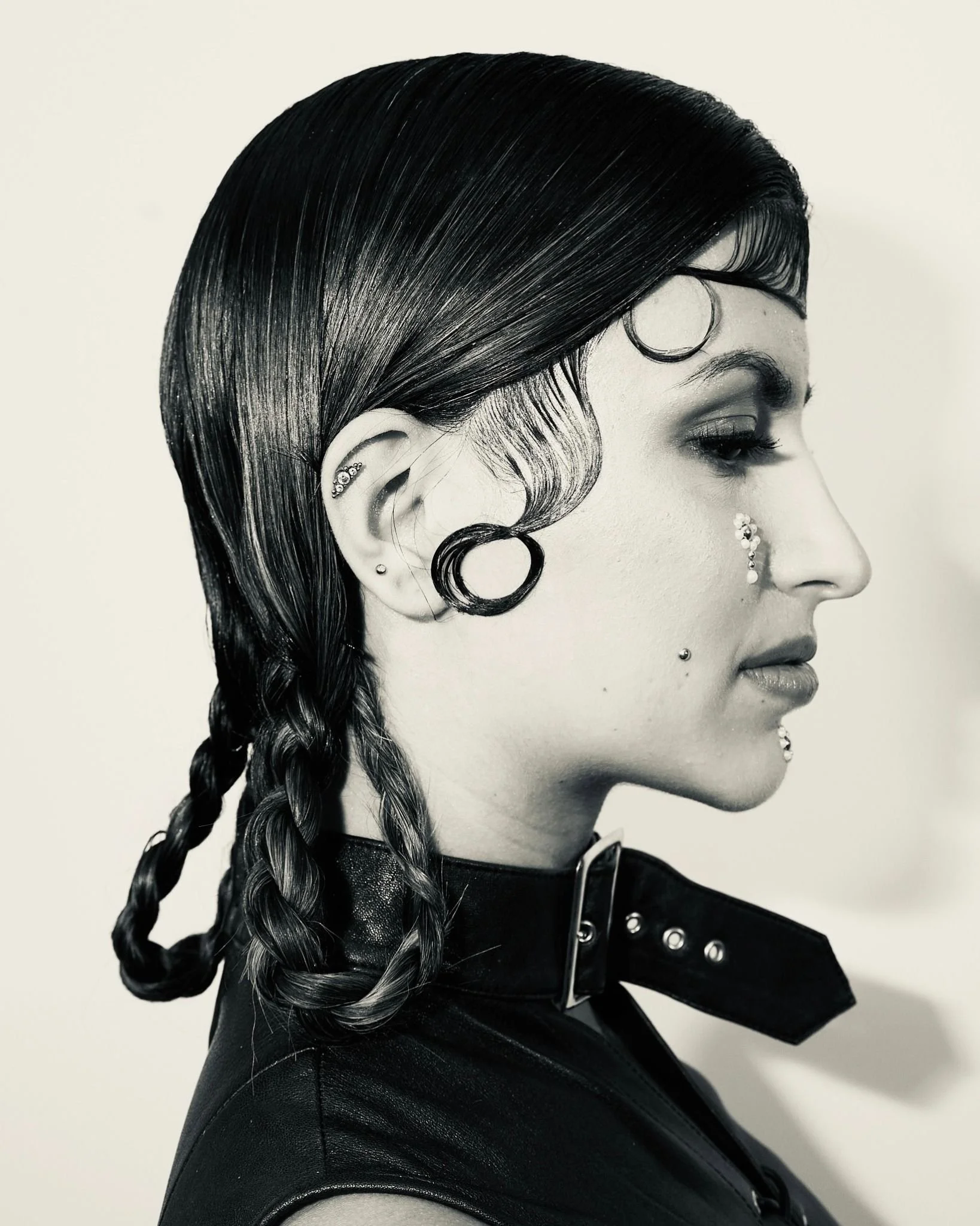Side profile of a woman with long, shiny black hair styled in a braid. She has multiple earrings and is dressed in a black leather jacket.