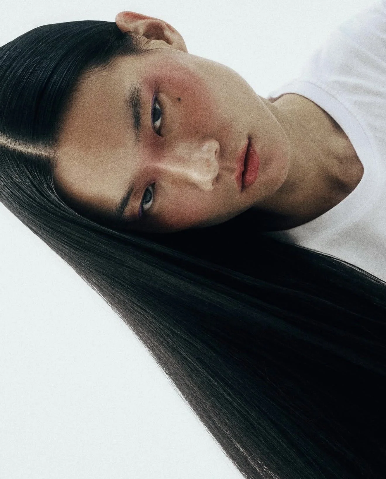 Close-up of a woman with long black hair, right eye winking, and wearing a white shirt against a plain background.