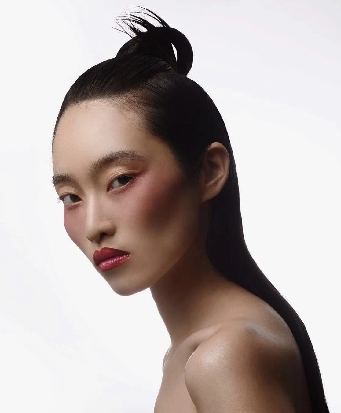 A portrait of a woman with sleek black hair styled in an updo, wearing makeup with red lipstick and blush, against a plain white background.