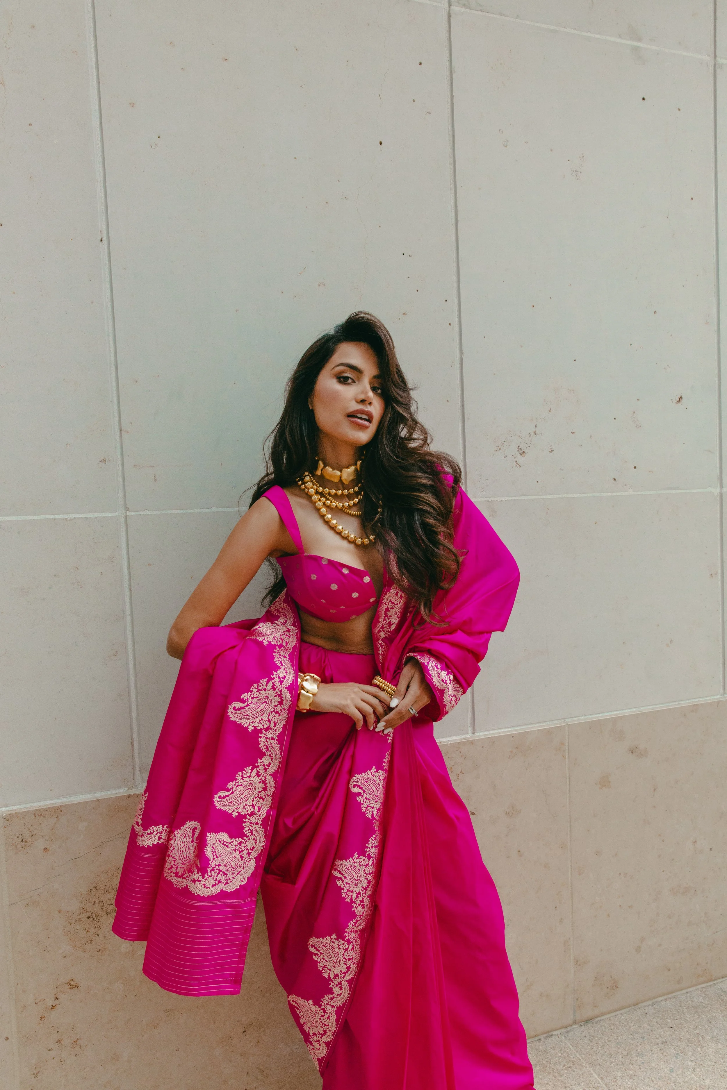 Woman in traditional bright pink Indian saree with gold jewelry, standing against a plain beige tiled wall.