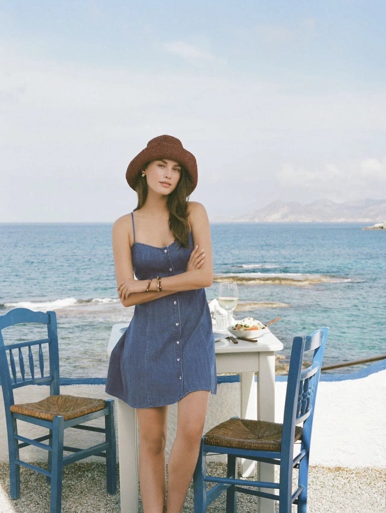 A young woman in a blue dress and a wide-brimmed hat stands by a white dining table with a seafood dish and a glass of white wine, with the ocean and distant mountains in the background.