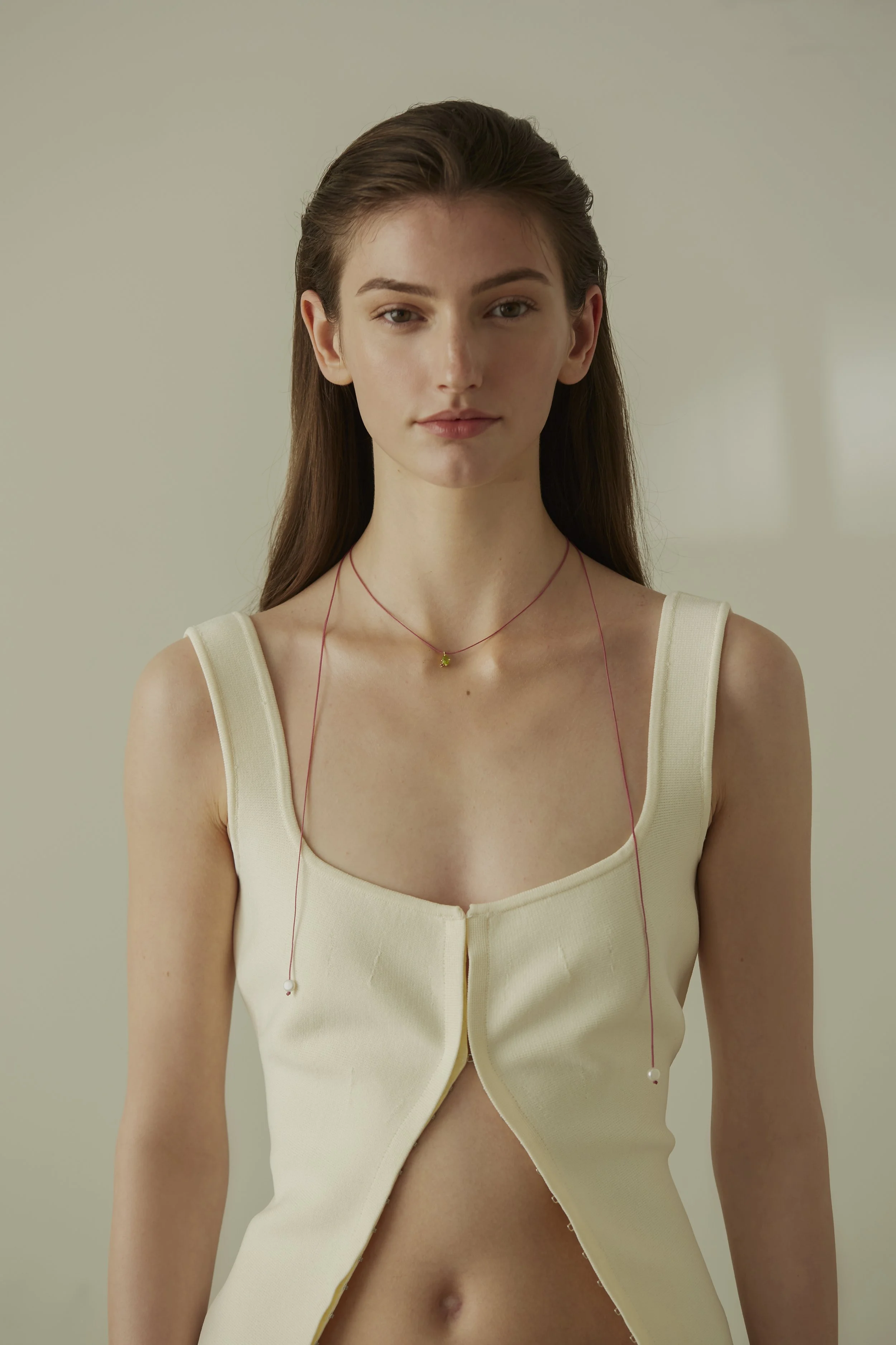 A young woman wearing a cream-colored top with her hair styled back and pink earphones around her neck, standing against a plain light beige background.