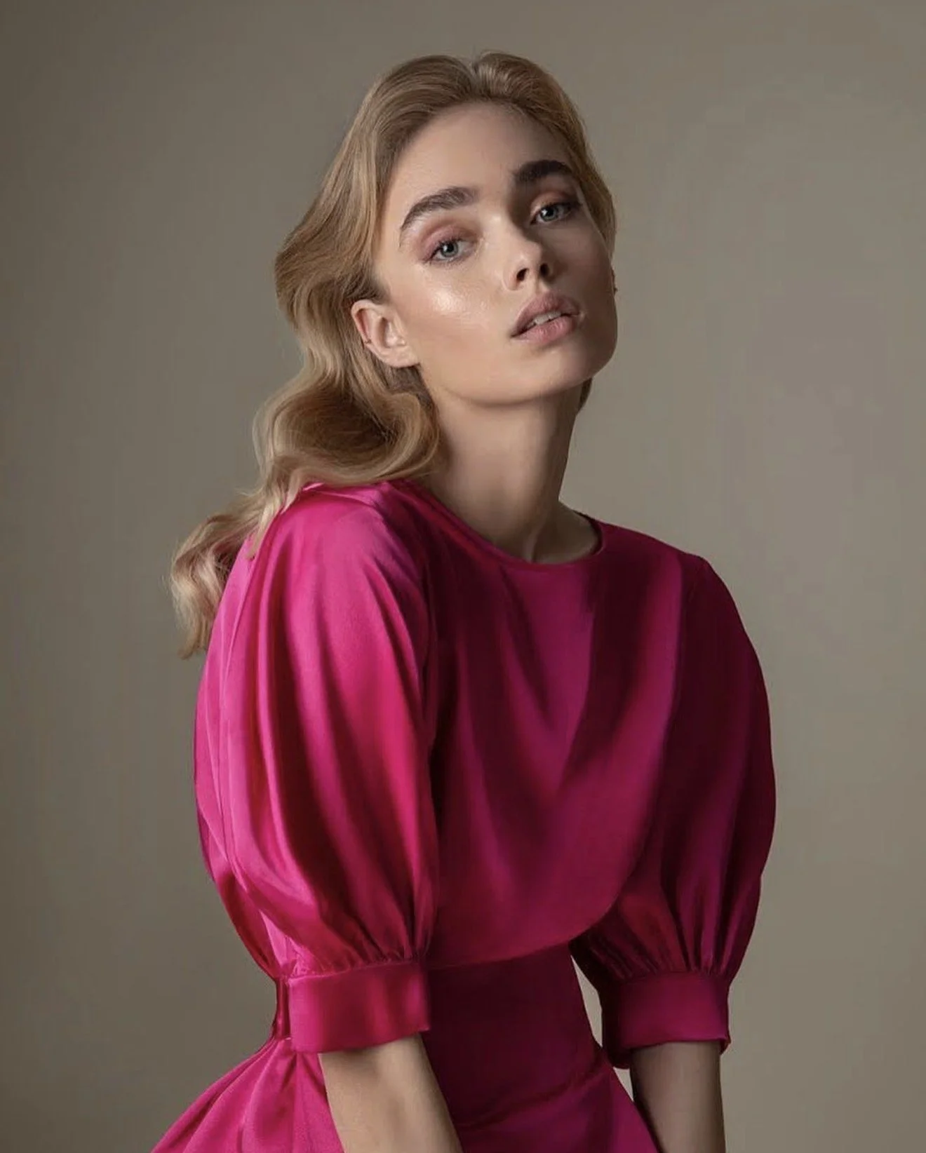 A woman with light skin and wavy blonde hair wearing a vibrant pink dress with puffed sleeves, looking at the camera with a neutral expression, against a plain gray background.