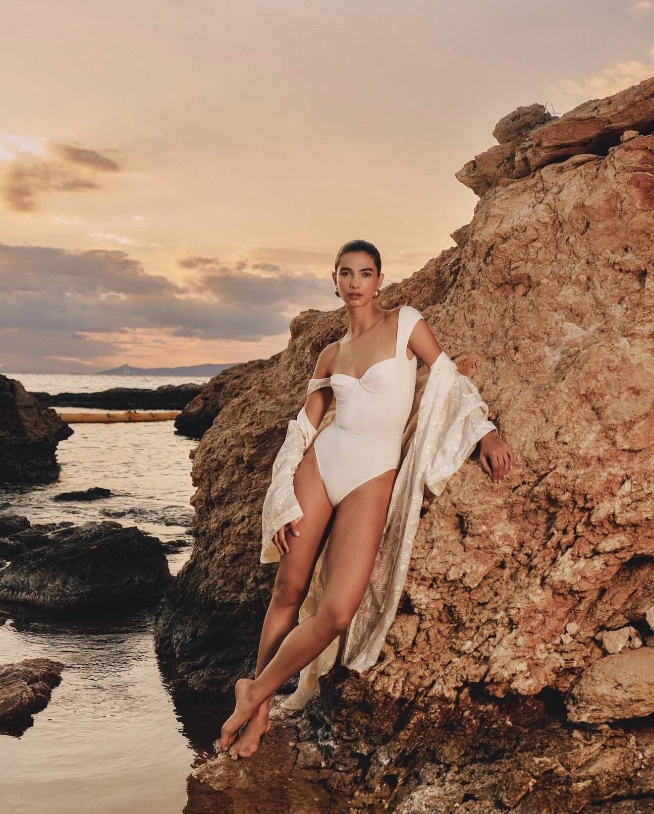 A woman in a white bodysuit and a sheer shawl leans against a large rock formation by the ocean at sunset.