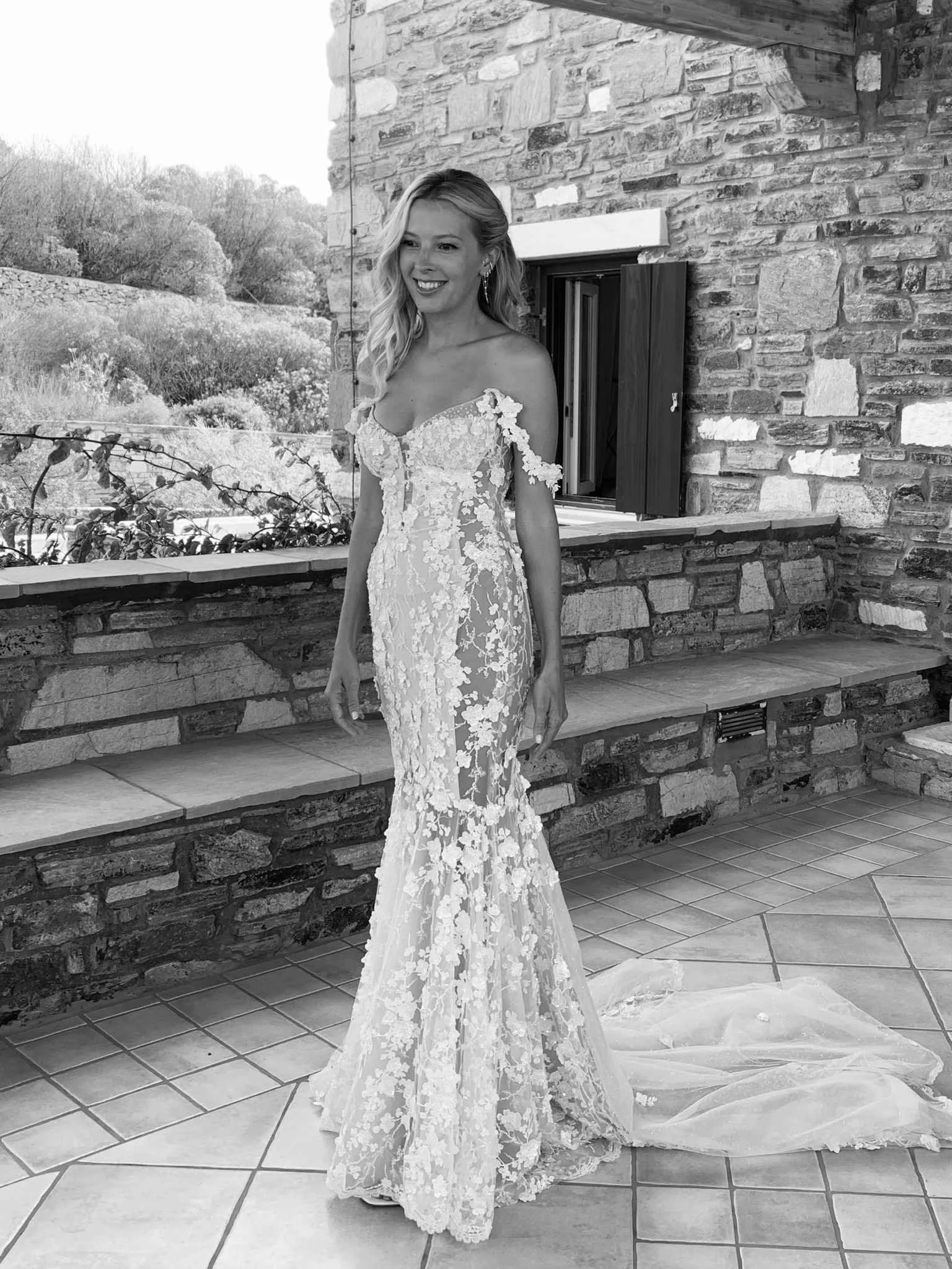 Black and white photo of a woman in a lace wedding gown standing outdoors near a brick wall and window, smiling.