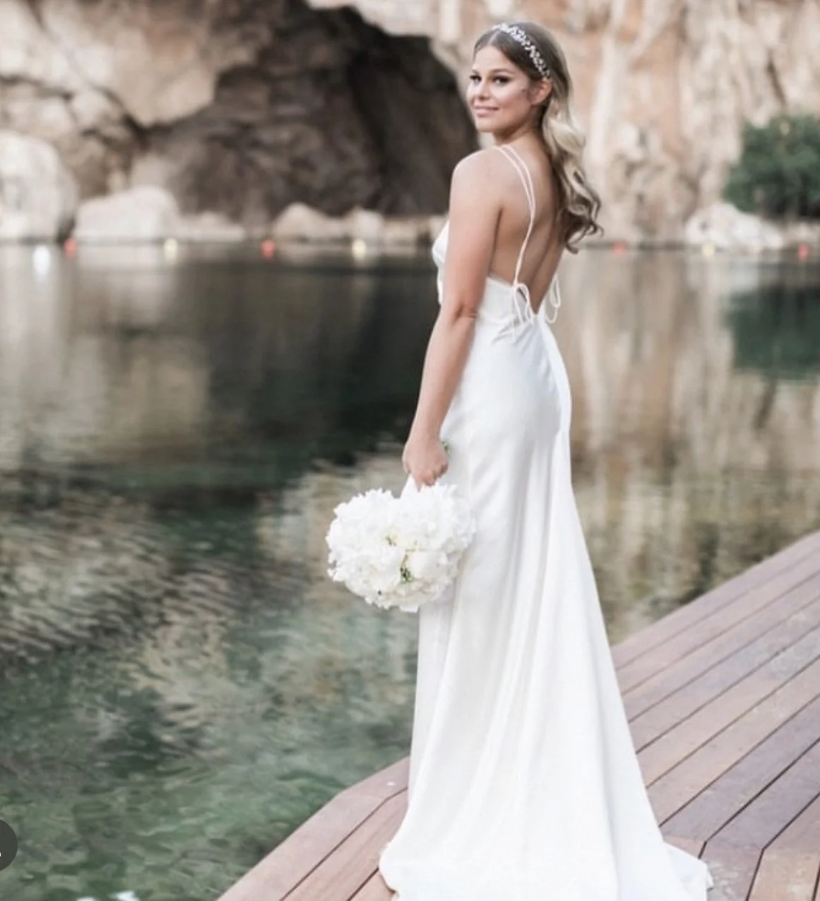 A bride in a white wedding dress holding a bouquet of white flowers, standing on a wooden dock by the water, with rocky hills in the background.