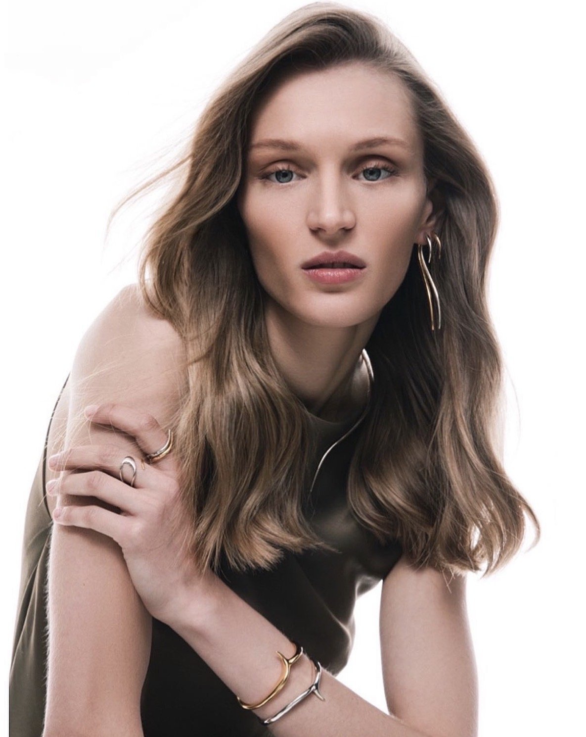A young woman with long, wavy brown hair and blue eyes, wearing earrings and bracelets, looking at the camera with a neutral expression against a white background.
