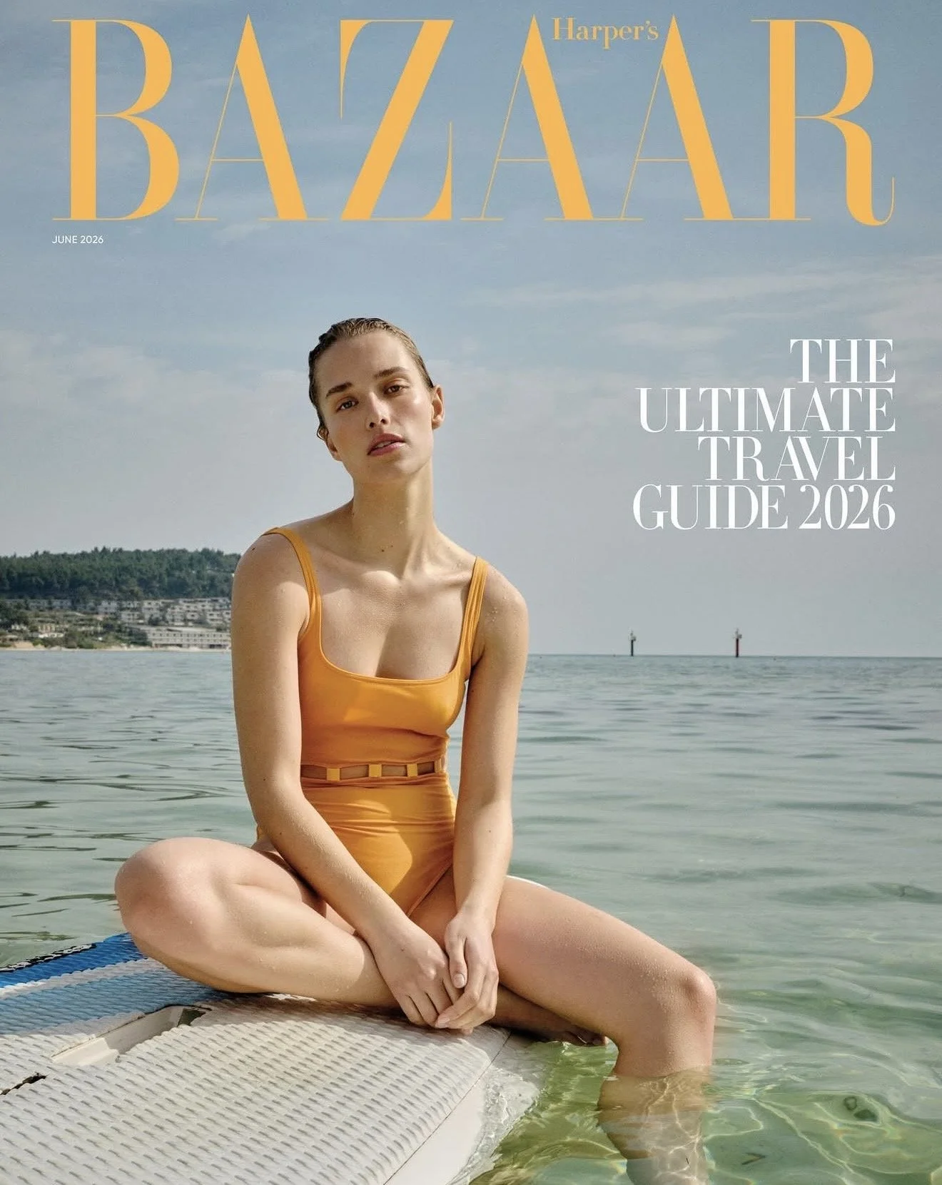 Cover of Harper's Bazaar June 2026 issue featuring a young woman in a yellow swimsuit sitting on a dock by a body of water with a cityscape and a cloudy sky in the background.
