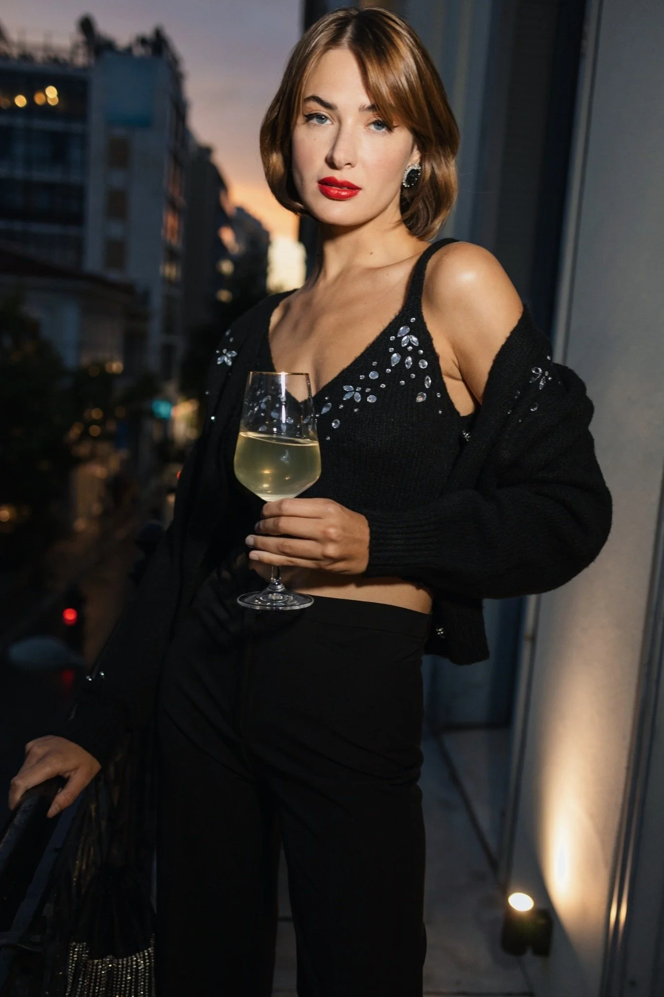A woman with short brown hair and red lipstick holding a glass of white wine, wearing a black top with rhinestone embellishments and a black cardigan, standing on a balcony during sunset with city buildings in the background.