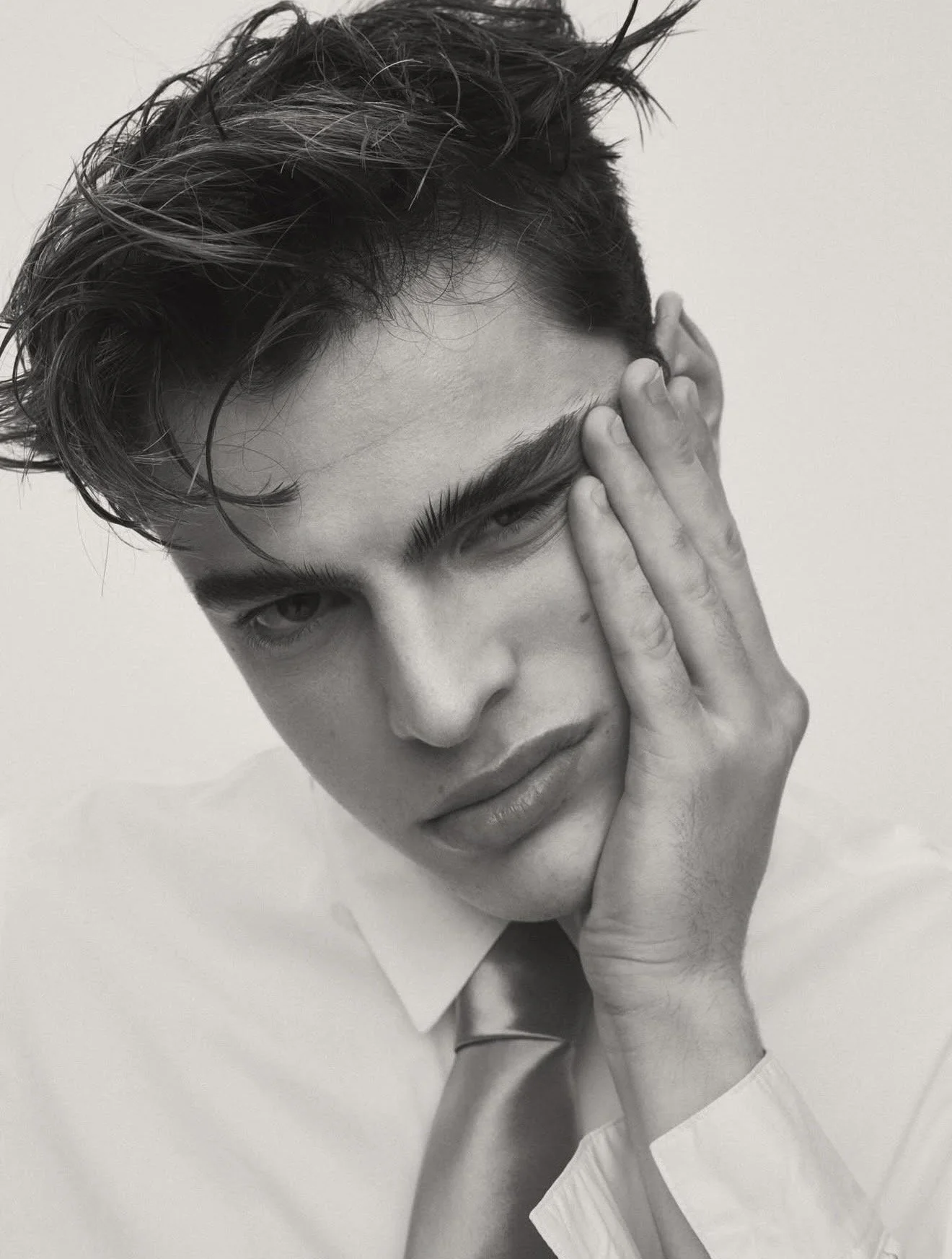 Black-and-white portrait photograph of a young person with wet, tousled hair, partially covering one eye, touching their face with one hand, wearing a collared shirt.