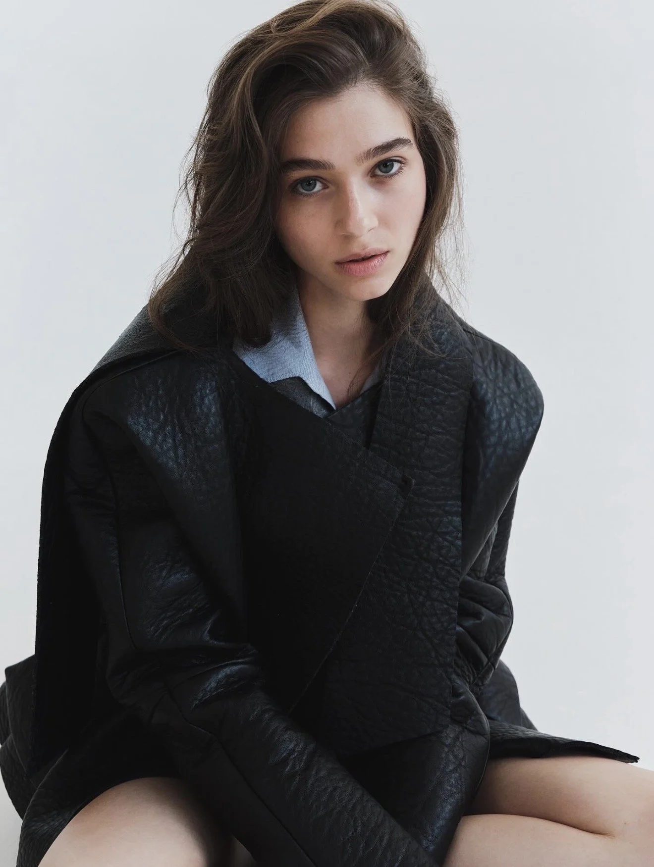 A young woman with long, dark brown hair and blue eyes looks directly at the camera. She wears a black textured jacket over a light blue shirt and sits with her right arm resting on her lap. The background is plain and light-colored.
